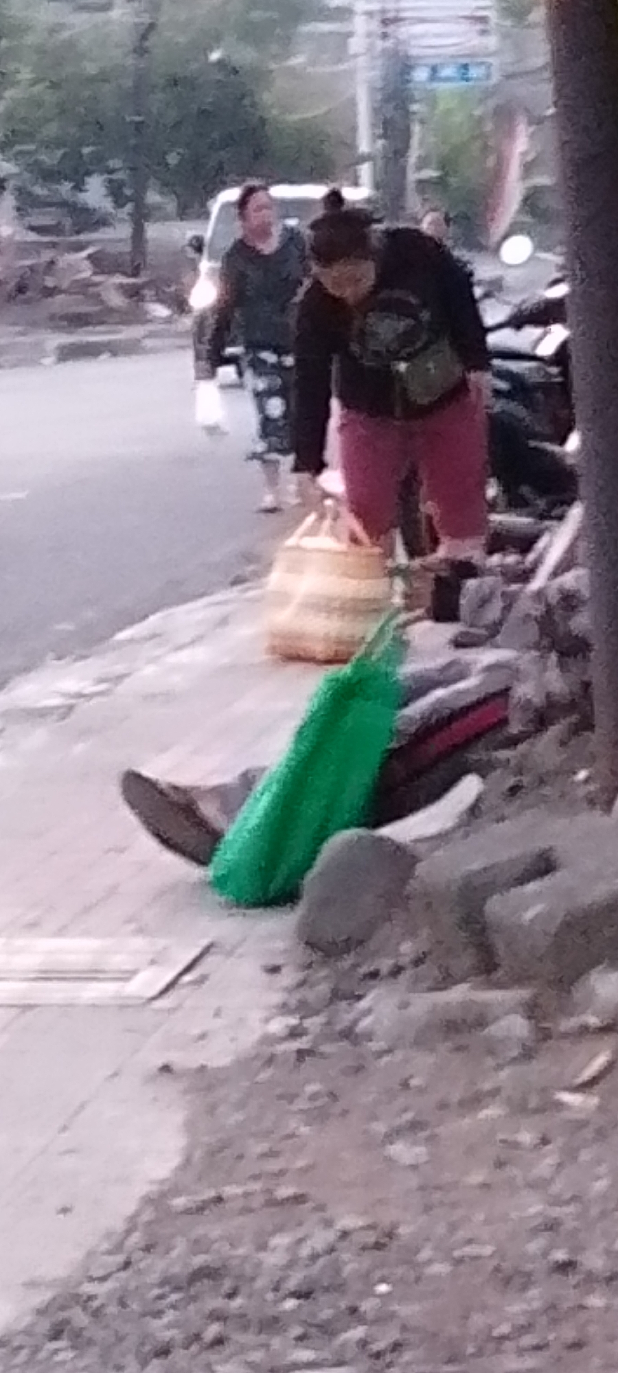 In Pekutatan, Indonesia, a person is seen sitting on the ground next to a pile of debris and rocks. They are wearing pink shorts and a black shirt, and a green bag is placed on their lap. A woven basket is on the ground nearby. In the background, several other individuals are walking on a paved surface, with a vehicle and trees visible.