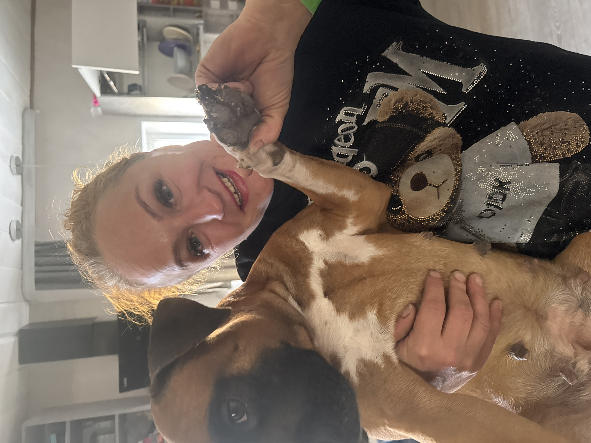 A woman indoors, with fair hair and red lipstick, holds a dog in her arms. She is smiling at the camera and holding up the dog's paw, which is covered in mud. The dog is mostly light brown with a dark brown face and white markings on its chest. The woman is wearing a black shirt with a teddy bear design. A kitchen with white cabinets is visible in the background. The location is not discernible from the image, and there is no indication it is in Chaikovsky, Russia.
