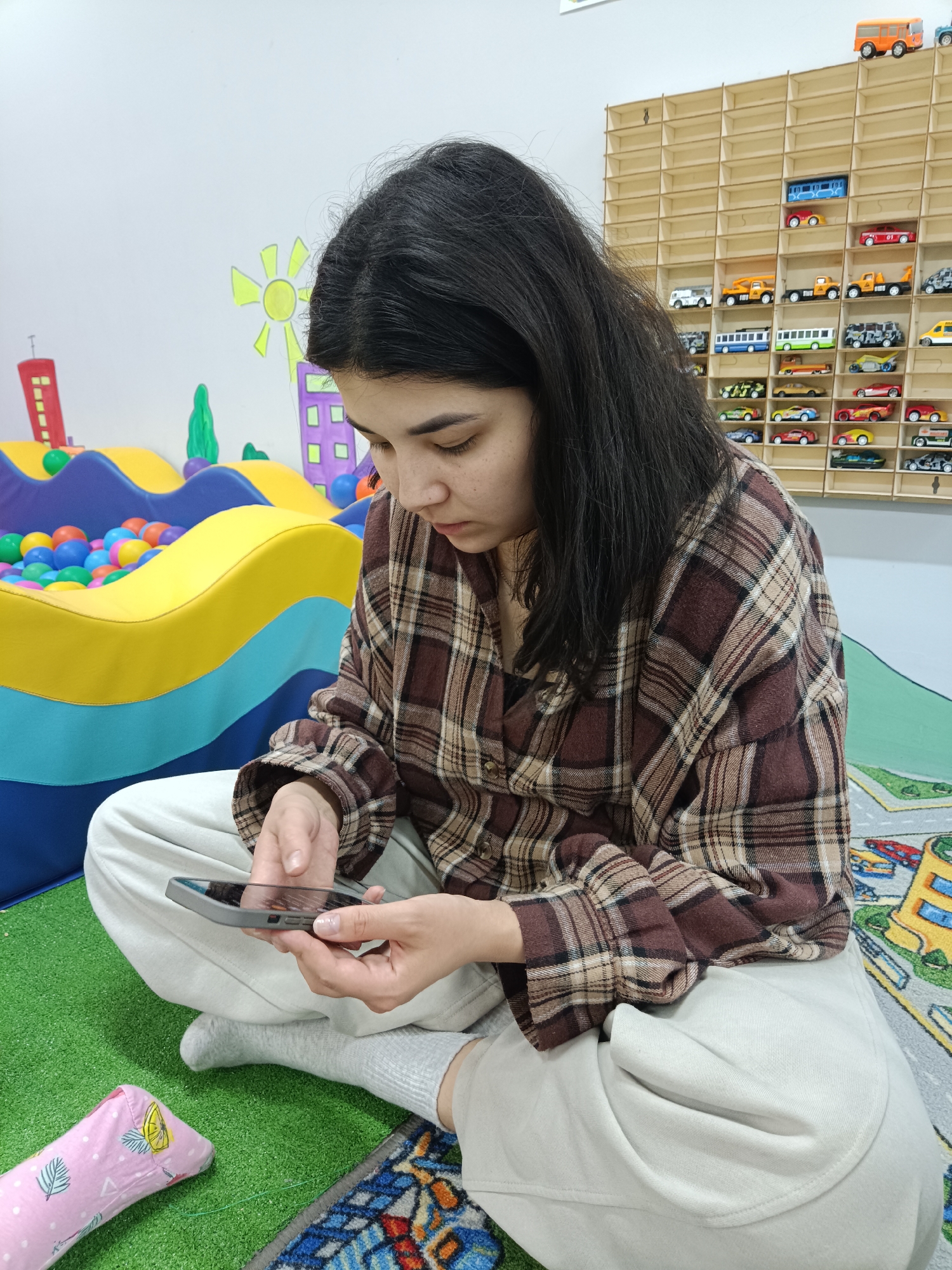 A young woman with dark hair is sitting cross-legged on a colorful carpet, holding a smartphone in her hands. She is wearing a brown and beige plaid shirt, light-colored pants, and gray socks. She appears to be focused on the screen of the phone. Behind her, there is a play area with a ball pit and a wall decorated with cartoon buildings and a sun. To her right, there is a wooden shelf filled with miniature toy cars. The overall environment appears to be a playroom or a children's activity area.