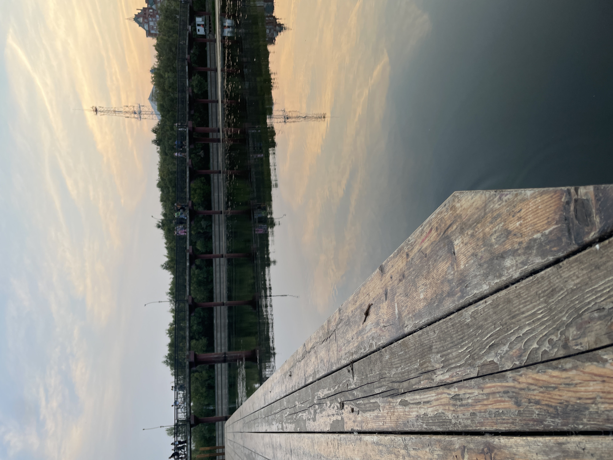 The image depicts a waterside scene at dusk, likely in Surgut, Russia. The perspective is from a wooden pier, with weathered planks filling the foreground. Beyond the pier, a calm body of water reflects the sky and the opposite bank. The sky is filled with wispy clouds tinged with the warm colors of sunset, suggesting the golden hour.
On the opposite bank, a line of trees forms a green backdrop. A bridge with reddish-brown support pillars spans the water. On the bridge, several small figures of people are visible, appearing to be strolling. In the background, behind the trees and bridge, are buildings and a tall radio tower. The reflection in the water clearly mirrors these elements, including the sky, trees, bridge, buildings, and the radio tower, creating a symmetrical visual effect. The water itself appears dark and still, with subtle ripples indicating a gentle breeze. The overall mood is peaceful and serene, characteristic of a quiet evening by the water. No visible text is present in the image.