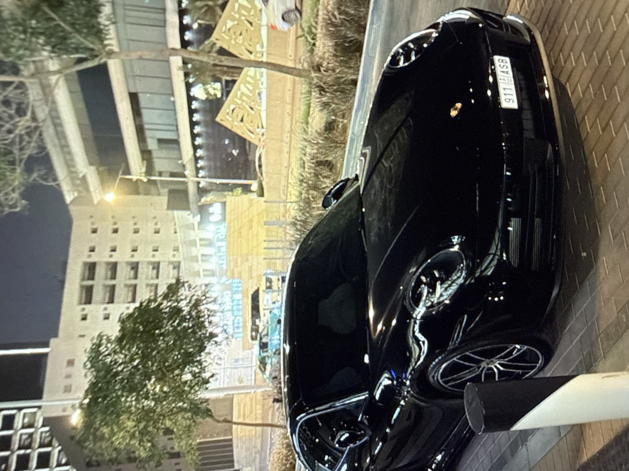 A black Porsche 911 is parked in the foreground, angled slightly towards the right, on a paved surface that appears to be a sidewalk or an adjacent parking area. The vehicle is highly reflective, showing various light sources and subtle architectural reflections. Its front-mounted license plate displays "911 ASB" with a blue strip on the left containing the EU flag and the country code "BG," confirming it as a Bulgarian registration. A black and white bollard is positioned in the immediate left foreground.
The scene is set at night, illuminated by streetlights and building lights. In the mid-ground, a multi-lane road is visible, with other vehicles in the distance, including a light-colored SUV on the far right, a light blue/green sedan, and a yellow/beige car. A median strip separates the road, featuring sparse, dry vegetation.
The background consists of several buildings. To the left, a multi-story, light-colored building with numerous rectangular windows is visible behind a green-leafed tree. To the right, a more modern, multi-story structure with extensive glass facades and a prominent overhang is illuminated by a bright streetlight. Further right, two large, angular, decorative panels with intricate cutout patterns flank the road, suggesting a distinctive architectural or public art element. These panels are illuminated, highlighting their geometric design. The environment indicates an urban setting within the city of Kardzhali, Bulgaria. No individuals are discernible in the image.