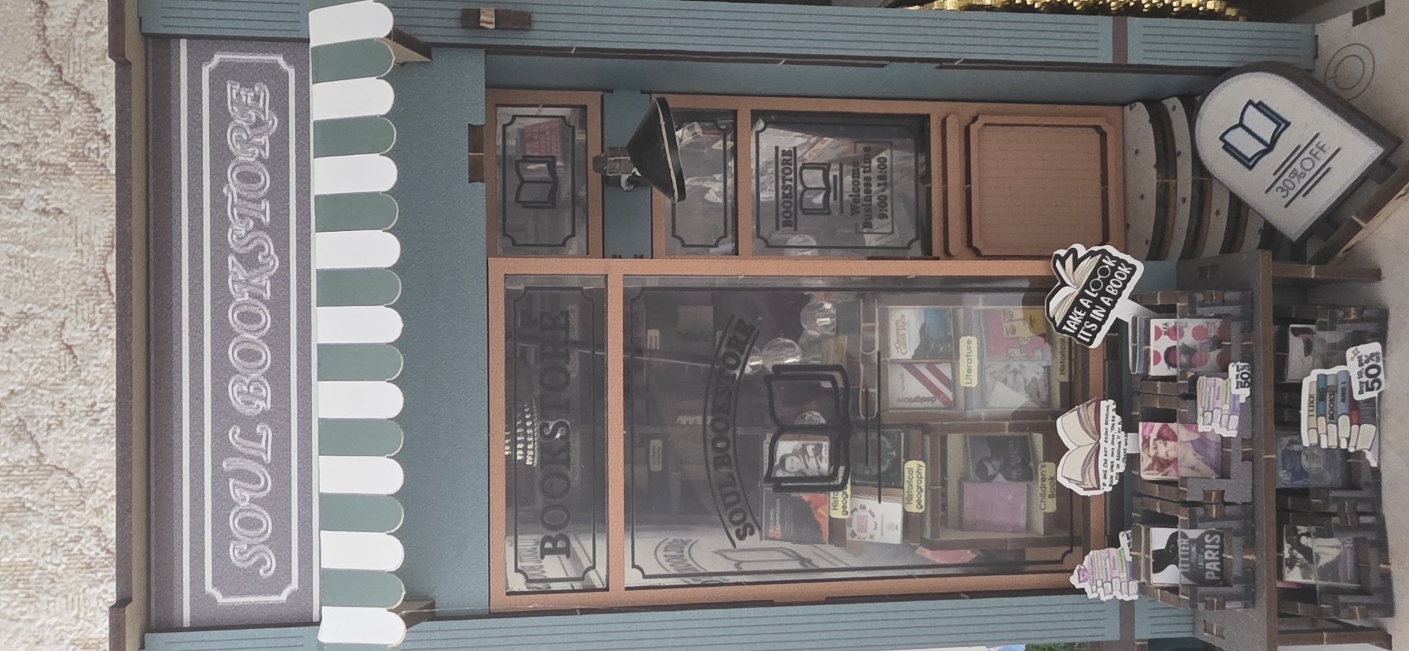 This is a close-up, high-angle, vertical shot of a miniature diorama depicting a bookstore. The diorama is made of wood and paper and is richly detailed.
**Subjects:** The primary subjects are the miniature books and shelves that make up the bookstore's interior. There are also decorative elements like a small lamp, signage, and a small sign advertising a 30% discount. A sign on the left side of the diorama reads "SOUL BOOKSTORE" in an ornate font. Inside the store, various books are displayed on shelves, with some appearing to have colorful covers.
**Objects:** Key objects include numerous miniature books of varying sizes and colors, wooden shelves, a display sign, a lamp, and decorative elements such as a stylized open book with text. There are also smaller signs with text like "BOOKSTORE", "Welcome Business time 9:00-18:00", "30%OFF", "TAKE A LOOK IT'S IN A BOOK", and "Buy 10, get 50%". Some books have titles such as "Literature", "Historical geography", "Children's Book", and "LETTER PARIS".
**Scene/Setting:** The scene is a meticulously crafted miniature bookstore. The exterior features a teal facade with white and green scalloped trim, mimicking an awning. The interior is designed to resemble a cozy, traditional bookstore with brick-patterned walls and wooden shelves stocked with books. The background outside the diorama appears to be a textured, light-colored surface.
**Activity/Event:** The diorama is a static representation of a bookstore, so no specific activity or event is taking place. It's a display of a place where books are sold and enjoyed.
**Notable Details:** The detail in the miniature books, including visible titles and illustrations on some covers, is striking. The signage adds to the realism and provides information about the bookstore, such as its name, hours, and promotions. The overall craftsmanship suggests this is a hobbyist creation or a model for a larger project. The lighting suggests it might be an indoor scene, but the specific time of day or weather cannot be determined from the diorama itself. No people are visible in the diorama.
**Visible Text:**
- SOUL BOOKSTORE
- BOOKSTORE
- Welcome Business time 9:00-18:00
- 30%OFF
- TAKE A LOOK IT'S IN A BOOK
- Historical
- Geography
- Literature
- Children's Book
- LETTER PARIS
- Buy 10, get 50%
- I LIKE BIG BOOKS AND I
The location context provided is Sumgayit, Azerbaijan. However, there are no visual cues within the image to confirm this specific location.
