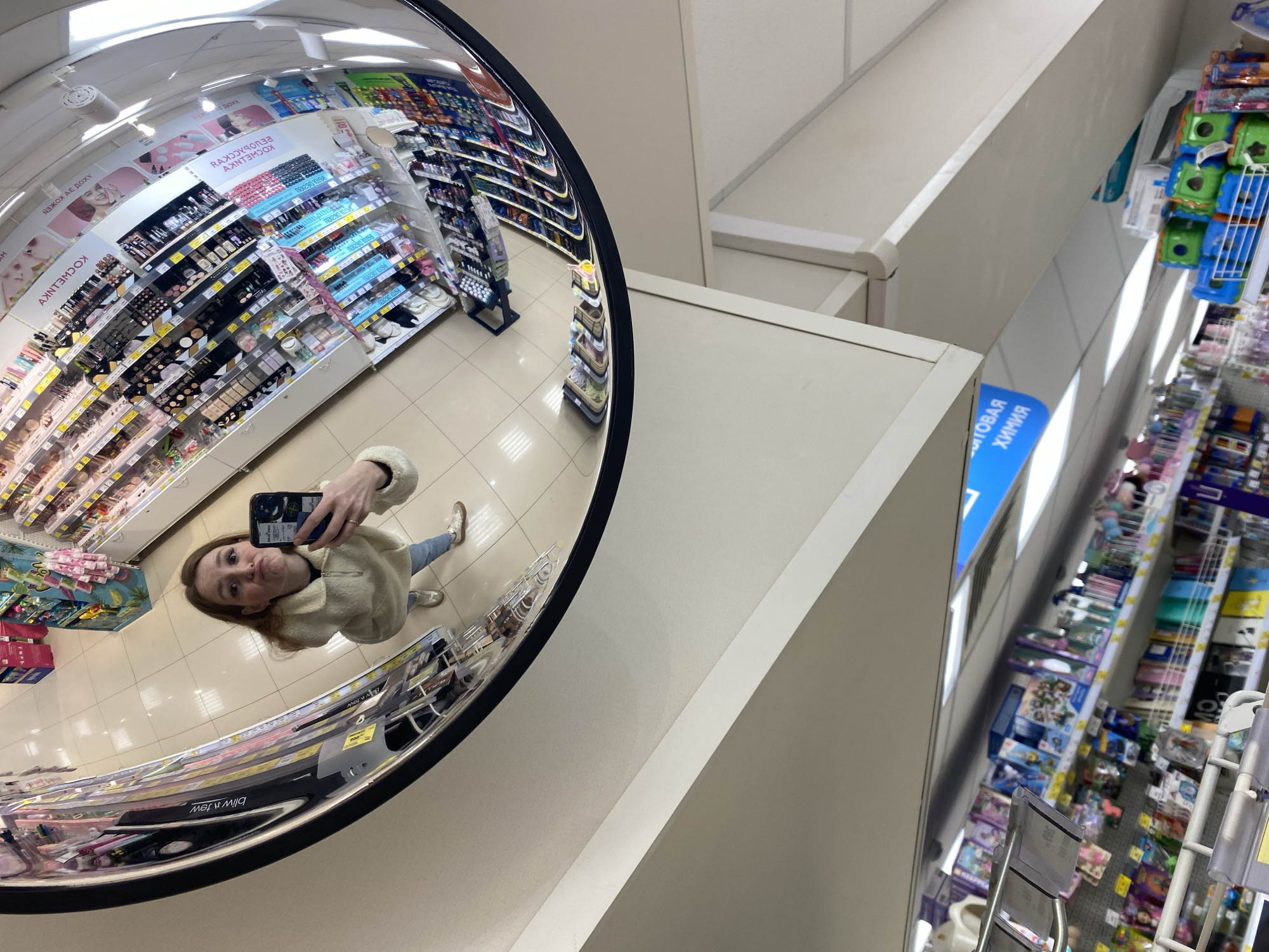 The image captures a selfie taken by a young woman in a large, black-framed convex security mirror inside a retail store. The woman, with reddish-brown hair, is wearing a light-colored, possibly cream, fluffy jacket, blue jeans, and light-colored sneakers. She holds a dark-colored smartphone to take the photo, making a slight pouting or "duck face" expression.
The mirror's reflection, occupying the left side of the frame, shows a wide-angle, distorted view of the store's interior. Numerous aisles and shelves stocked with various products are visible. Directly to the right of the mirror, the store's interior is seen from a different perspective, with more shelves and products. The store has light-colored tiled flooring and a white ceiling with visible lighting fixtures. The walls and structural elements are also light-colored.
The shelves, both in reflection and direct view, display a wide array of goods. In the mirror's reflection, signs for "КОСМЕТИКА" (COSMETICS) and "БЕЛОРУССКАЯ" (BELARUSIAN, likely referring to Belarusian cosmetics) are visible, indicating a section dedicated to beauty products, including various makeup items like eyeshadow palettes, mascaras, and lipsticks. On a blue sign directly visible in the store, the word "ХИМИЯ" (CHEMICALS) is decipherable, suggesting a household chemicals or cleaning supplies section.
The activity is the woman taking a playful selfie while shopping or browsing. The time of day is not evident, but the indoor lighting suggests business hours. The setting is a general merchandise store, supermarket, or a large cosmetics/drugstore in Chaikovsky, Russia.