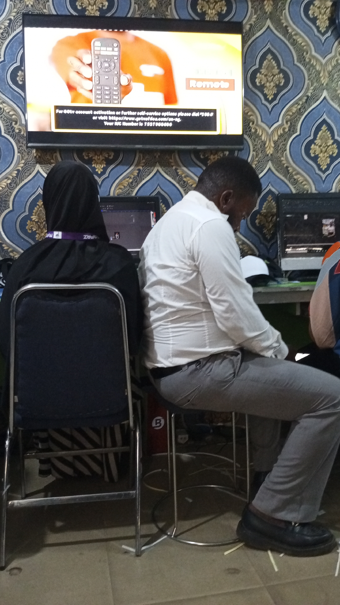 In an indoor setting, two individuals are seated at computers, facing away from the camera. The person on the left is wearing a black hijab and a purple lanyard, and is seated on a black chair with a metal frame. The person on the right is wearing a white button-down shirt and grey pants, and is seated on a stool. A large flat-screen television is mounted on a patterned wallpapered wall behind them, displaying an advertisement with a remote control and text in English. The environment appears to be a business or service center. The floor is tiled. The scene is consistent with an internet cafe or a similar establishment in Jos, Nigeria.