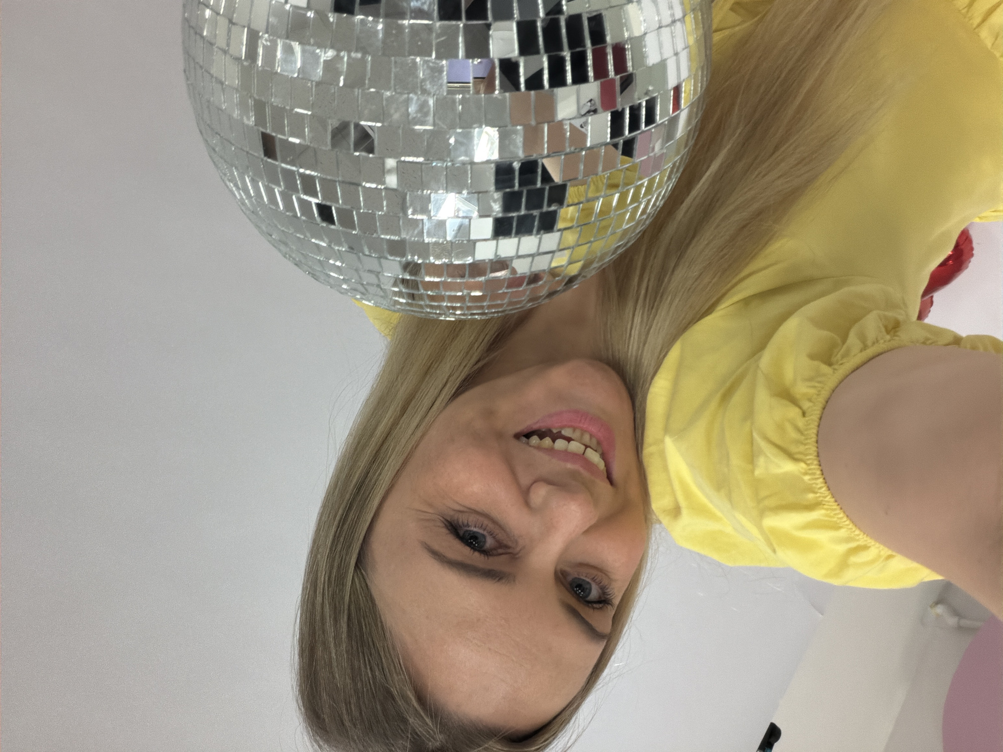 A selfie taken indoors from a low angle features a fair-skinned woman with blonde hair, blue eyes, and a yellow dress. She is positioned near a mirrored disco ball, slightly above her head. The woman is smiling, and her face is partially reflected in the disco ball. The background is a plain white wall. Other balloons can be seen in the background. There is no clear indication of a location in Votkinsk, Russia.