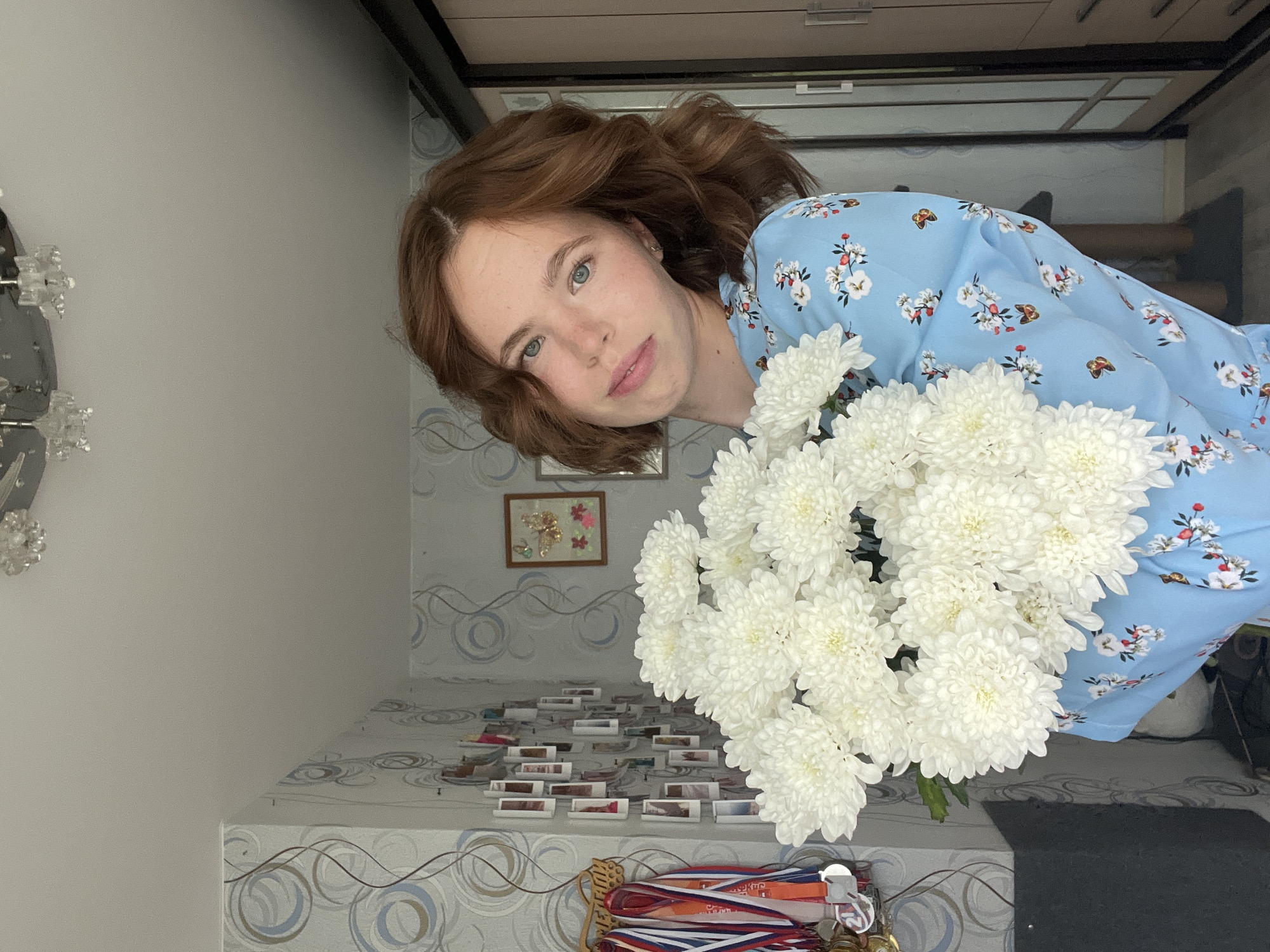 The image depicts a young woman with auburn hair and fair skin, holding a large bouquet of white chrysanthemums. She is wearing a light blue dress adorned with a floral pattern featuring red and white flowers and small brown butterflies. Her gaze is directed towards the camera, and she has a gentle smile.
The background suggests an indoor setting. To the left, a wall is covered in patterned wallpaper with circular designs in blue, grey, and brown. A collection of small framed photographs is displayed on this wall, alongside a wooden rack holding several medals on ribbons. In the upper left corner, a ceiling light fixture with crystal accents is visible. To the right, a portion of a wardrobe or closet with a beige finish is present. The floor appears to be a dark grey carpet or rug. A tall object, possibly a cat tree, is visible in the background to the right of the woman.
The overall scene is brightly lit, likely with natural light illuminating the room. There is no explicit indication of the city of Chaikovsky, Russia, within the image itself.