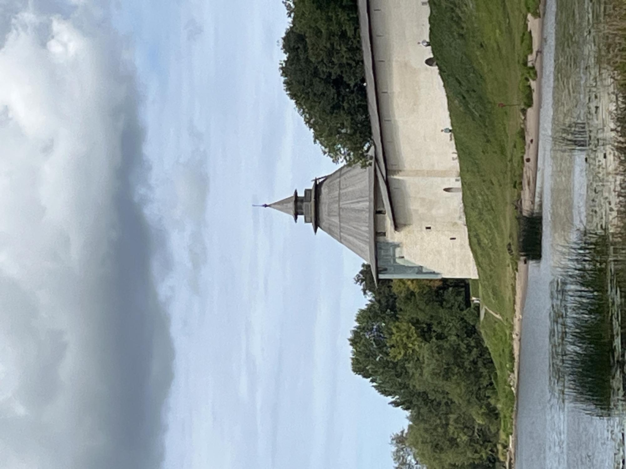 The image shows a view of the Pskov Kremlin in Pskov, Russia. A large, light-colored fortress wall with a conical-roofed tower is visible on a grassy hill overlooking a body of water. The sky is cloudy. Two small figures are visible walking on the grassy slope near the fortress wall. In the foreground, the water is partially covered with reeds and lily pads.