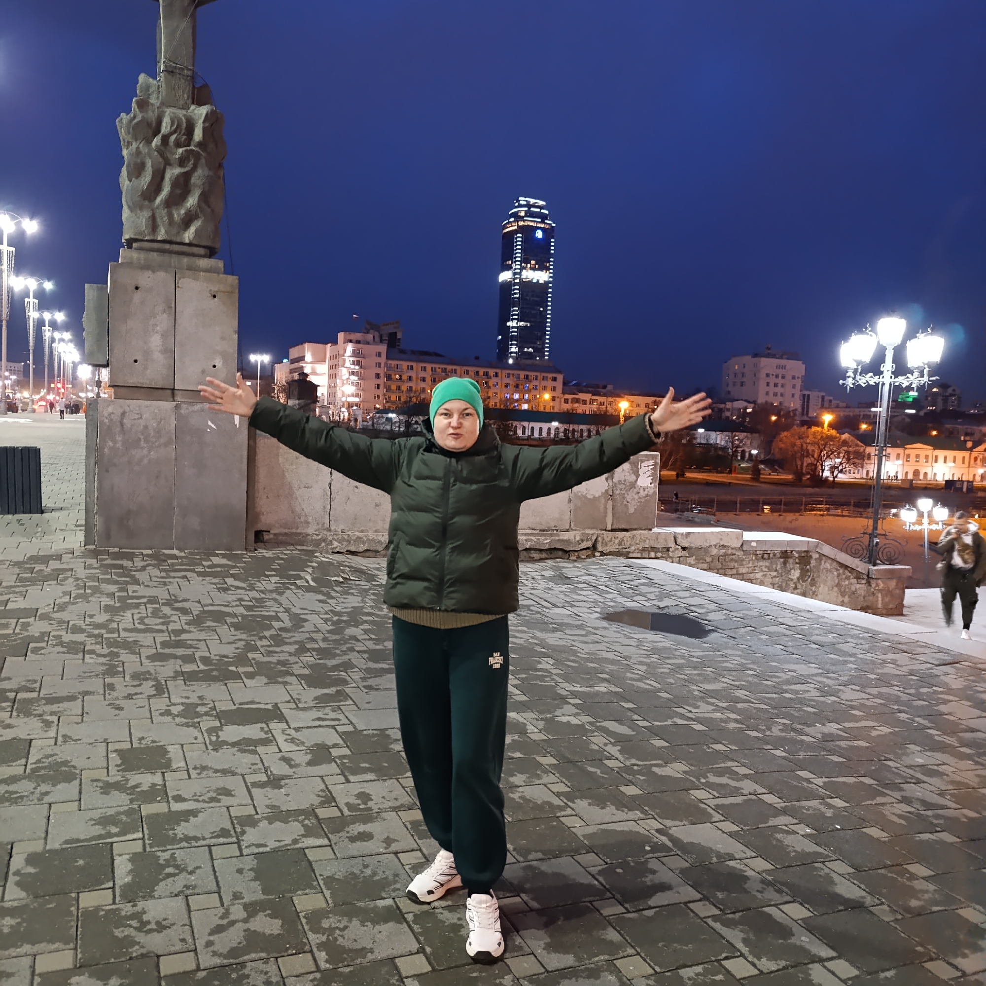 A woman stands with arms outstretched in Yekaterinburg, Russia. She wears a green puffy jacket, green sweatpants, a green knit cap, and white sneakers. She stands on a paved walkway with a statue and streetlights to her left. Behind her are various buildings, including a tall illuminated skyscraper. A person walks in the background to the right. The time of day is evening.