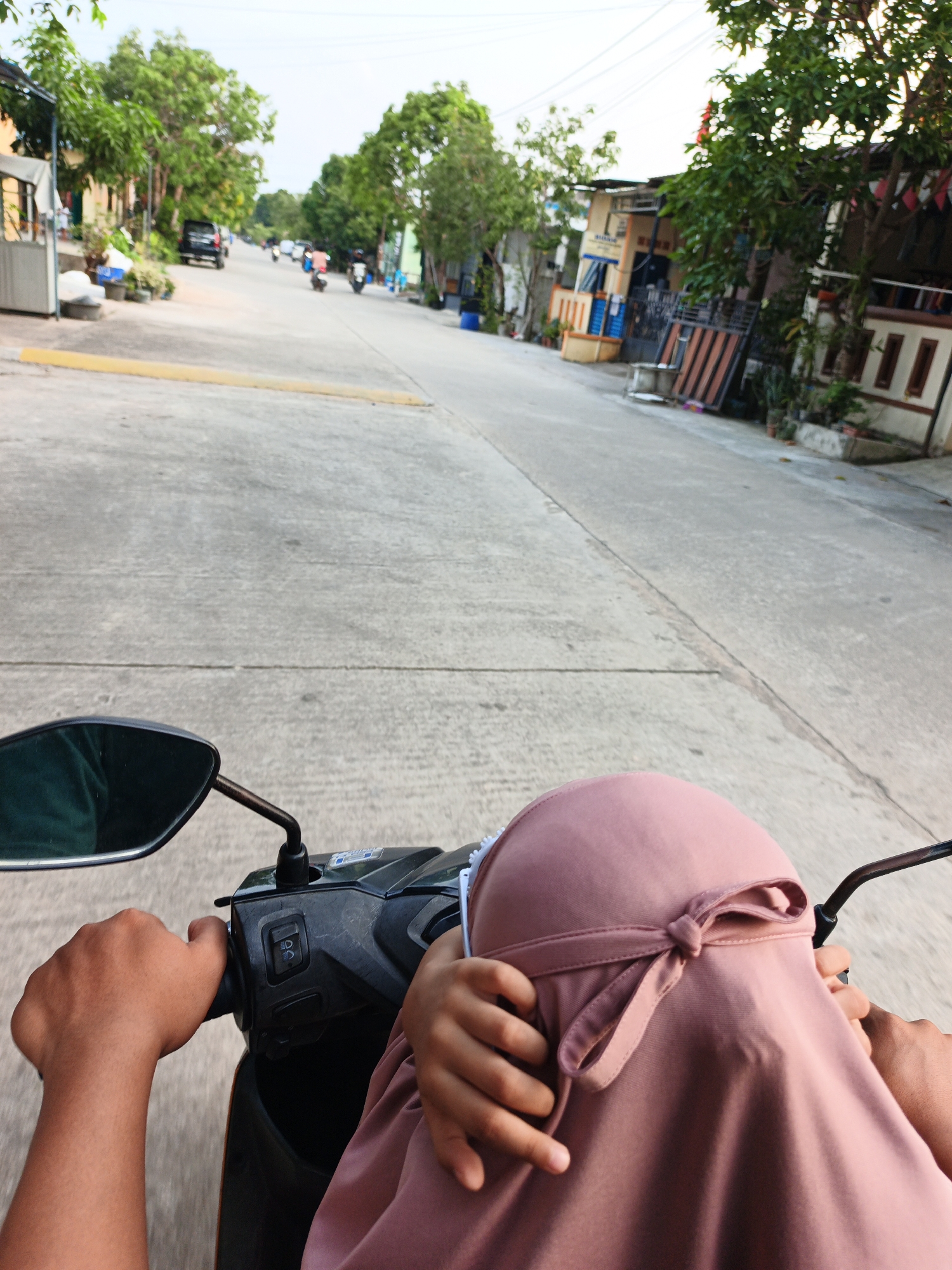 This is a first-person point-of-view photograph taken from the back of a motorcycle on a street in Batam, Indonesia. The road is made of concrete and has a yellow line running down its center. On the sides of the road are houses, trees, and a few parked vehicles. The sky is overcast. In the foreground, a person's hands are visible on the motorcycle's handlebars. A child, wearing a pink hijab tied with a ribbon, is seated in front of the rider, with their hands resting on the rider's arm. The image captures a common mode of transportation in the region.