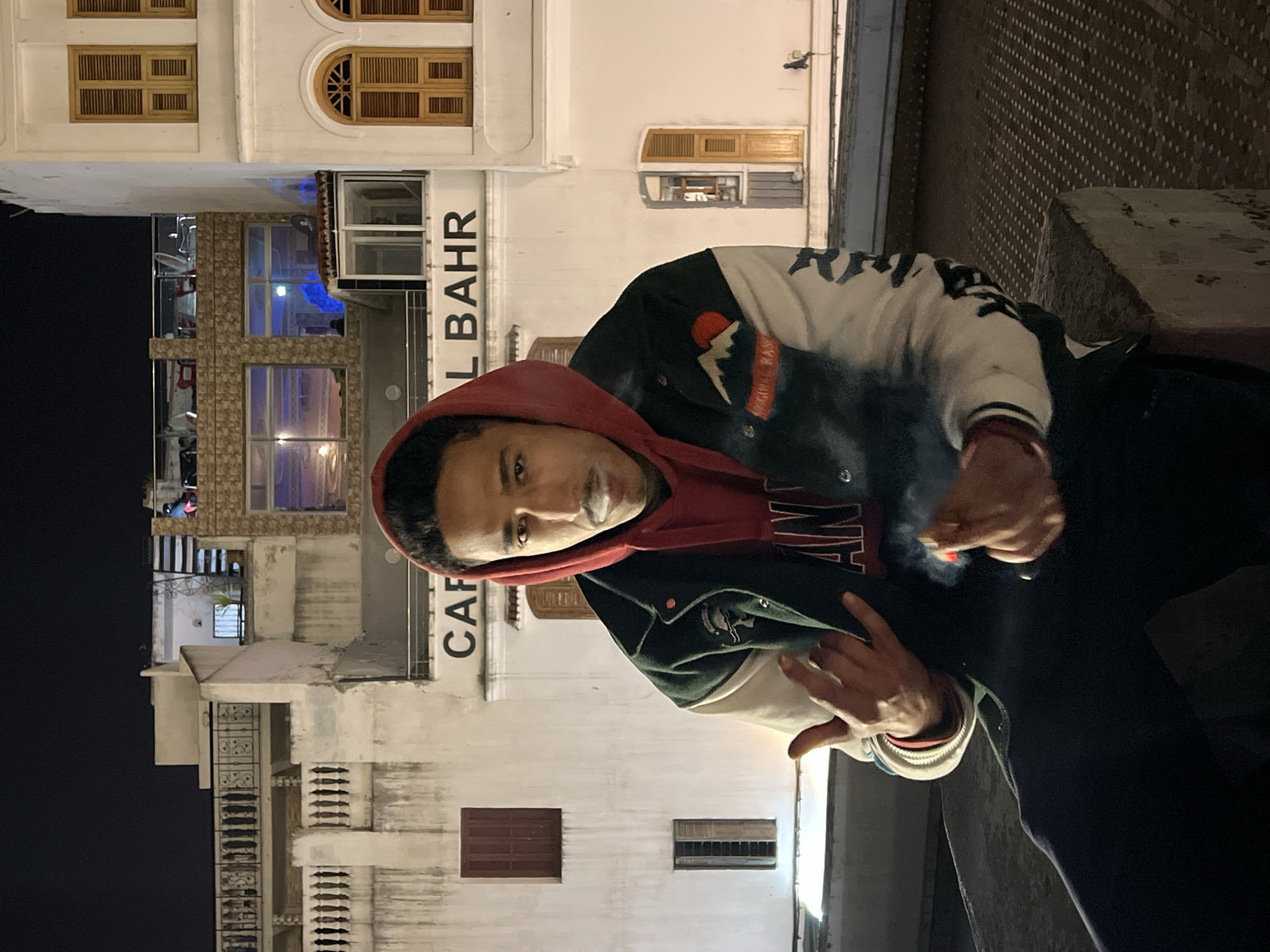 A male individual is depicted at night, seated on a low concrete barrier, looking directly at the camera. He wears a red hooded sweatshirt with the hood up, underneath a dark green and off-white varsity jacket featuring a mountain and sun patch on the right sleeve and partial text "ORIGINAL RAS". His left hand is raised in a "devil horns" gesture, while his right hand holds a lit cigarette, from which a plume of white smoke rises, partially obscuring his mouth and mustache. A red bracelet is on his left wrist.
The scene is an urban environment in Tanger طنجة, Morocco. In the background, multiple white-plastered buildings are visible under a dark sky, illuminated by artificial lighting. A prominent sign "CAFE AL BAHR" is inscribed on a building facade. Above this cafe, a rooftop terrace with visible patrons and tables reflects blue interior lighting. The architecture includes various windows, some with light wooden shutters, and decorative metal balcony railings. The ground surface in the foreground is dark and textured, likely paved.