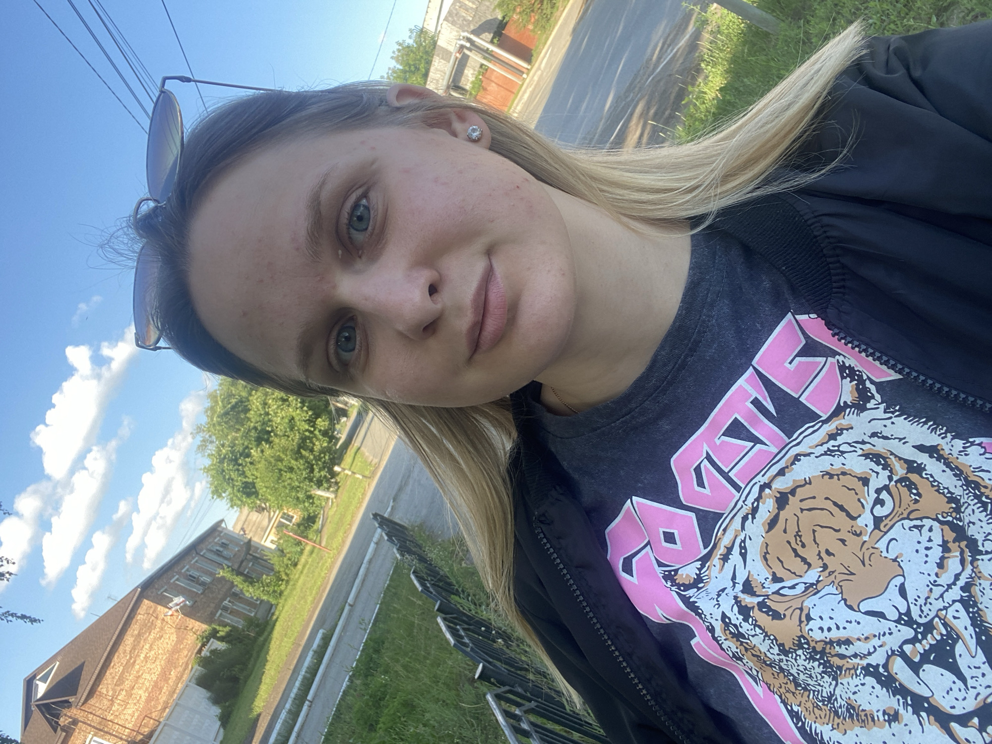 A young blonde woman is taking a selfie outdoors during the day. She is wearing a black jacket over a dark t-shirt with a large graphic of a tiger and pink lettering. She has light blue eyes, fair skin with some blemishes, and her blonde hair is visible around her face and shoulders. Sunglasses are perched on top of her head. The background shows a street with a grassy verge, a black metal fence, and buildings in the distance. The architecture and vegetation suggest a residential area in a city, consistent with Chaikovsky, Russia. The sky is blue with scattered clouds.