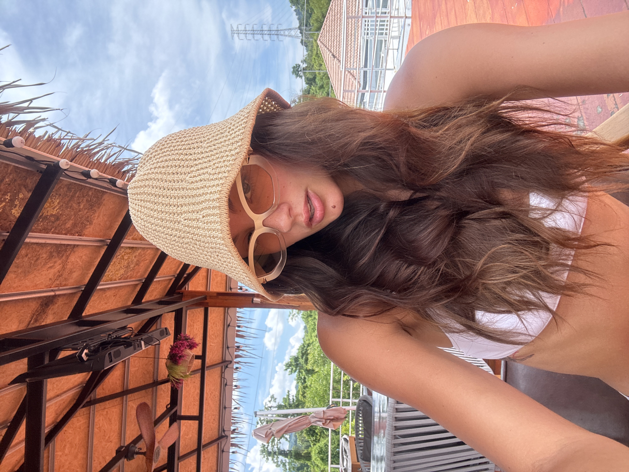 A woman in a white bikini and straw hat is posing in what appears to be a vacation setting in Kathu, Thailand. She is wearing sunglasses and is positioned under a thatch roof structure. In the background, there is a clear sky with some clouds, lush green trees, a roof of a building, and an electrical transmission tower. Part of a building with a metal structure is visible. There is also a fan attached to the ceiling.