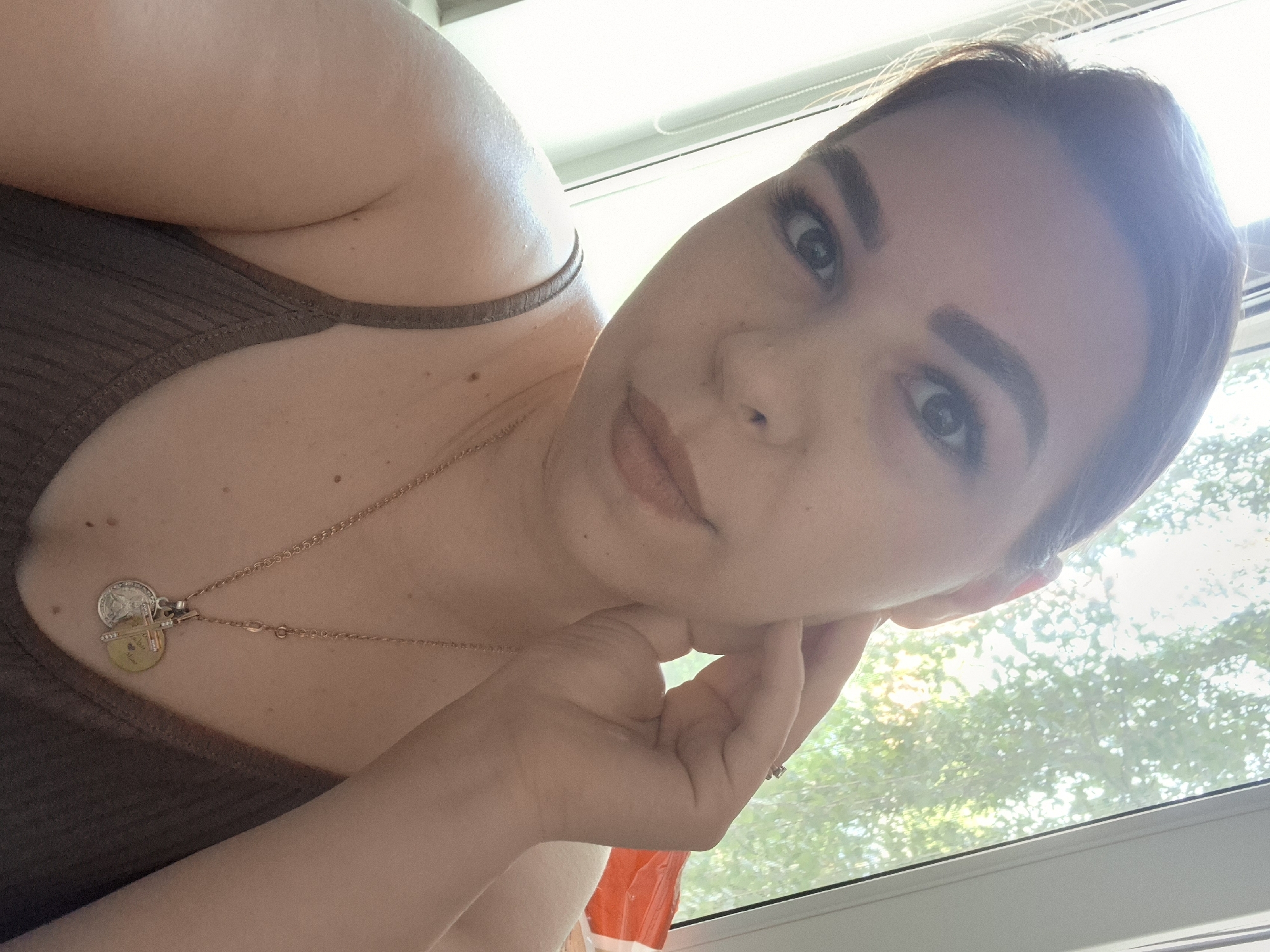 The image is a selfie taken indoors, with a window in the background showing greenery and sunlight. The subject is a woman with dark hair, wearing a brown ribbed tank top and a necklace with two pendants. Her right hand is touching her face. She is wearing subtle makeup, including mascara and lipstick. The overall lighting suggests it is daytime. There is no visible indication of the specific location within Chaikovsky, Russia.
