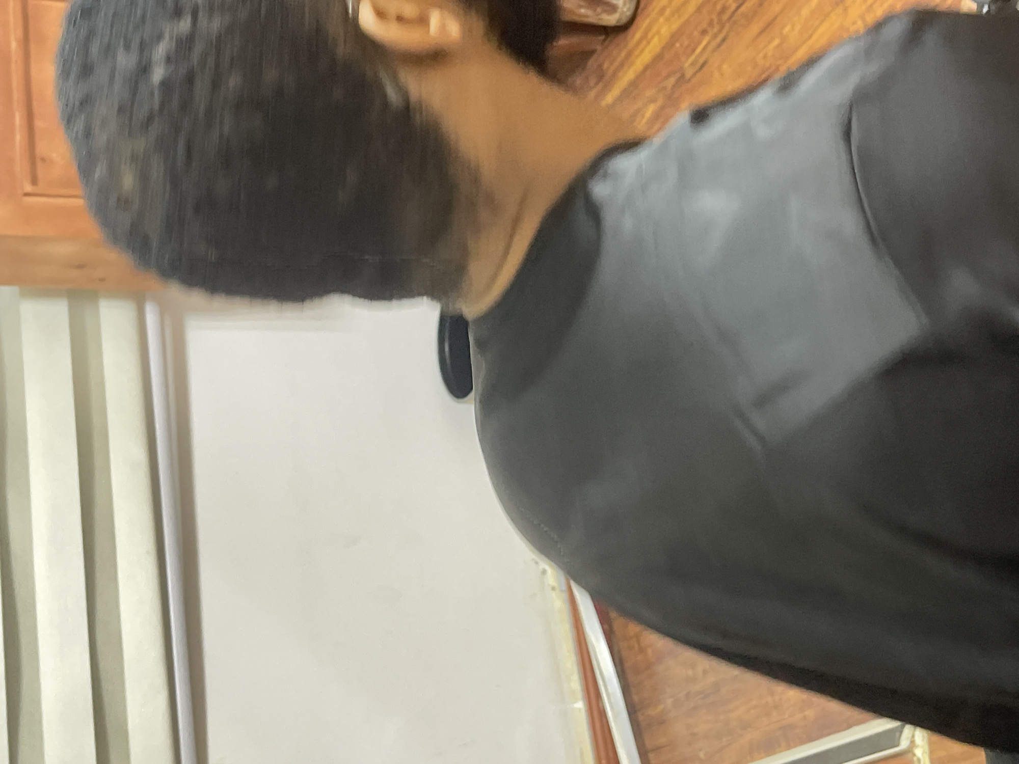A person wearing a black garment is positioned in front of a light-colored wall with vertical stripes. The wall is adjacent to a wooden surface. The person's head is turned away from the camera, showing the back of their head and neck. The location is implied to be Abuja, Nigeria.