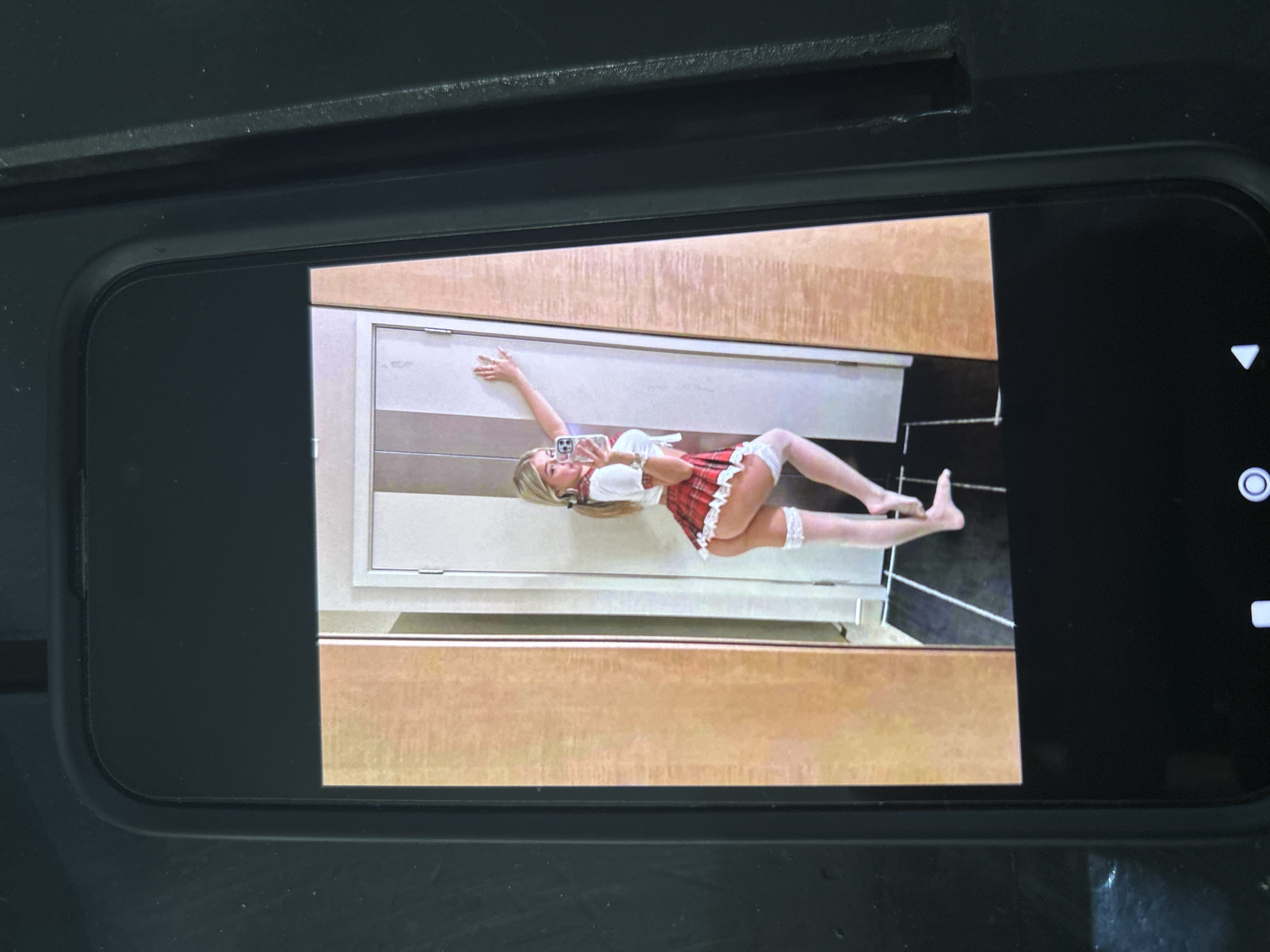 A vertically oriented photo of a phone screen displaying an image, resting on a dark surface. The phone screen shows a full-body picture of a person, possibly a woman, with fair skin and blonde hair, wearing what appears to be a schoolgirl-style outfit with a white top, a red and white plaid skirt, and white stockings or knee-high socks. She is lying on her back on a light-colored floor, with her head near the bottom of a doorframe or wall. Her left arm is extended upwards and bent at the elbow, with her hand resting against the wall. Her right arm is bent, and she is holding a smartphone up to take a selfie. Her legs are slightly bent.
The background of the displayed image includes a light-colored doorframe or wall, with a darker floor visible in the lower right portion of the image. Above the doorframe, a brown, wood-like surface is visible. The phone displaying the image has a black bezel and shows standard smartphone interface elements on the right side, such as a camera icon (a circle with a smaller circle inside) and possibly navigation arrows. The overall setting appears to be indoors. There is no visible text in the image.