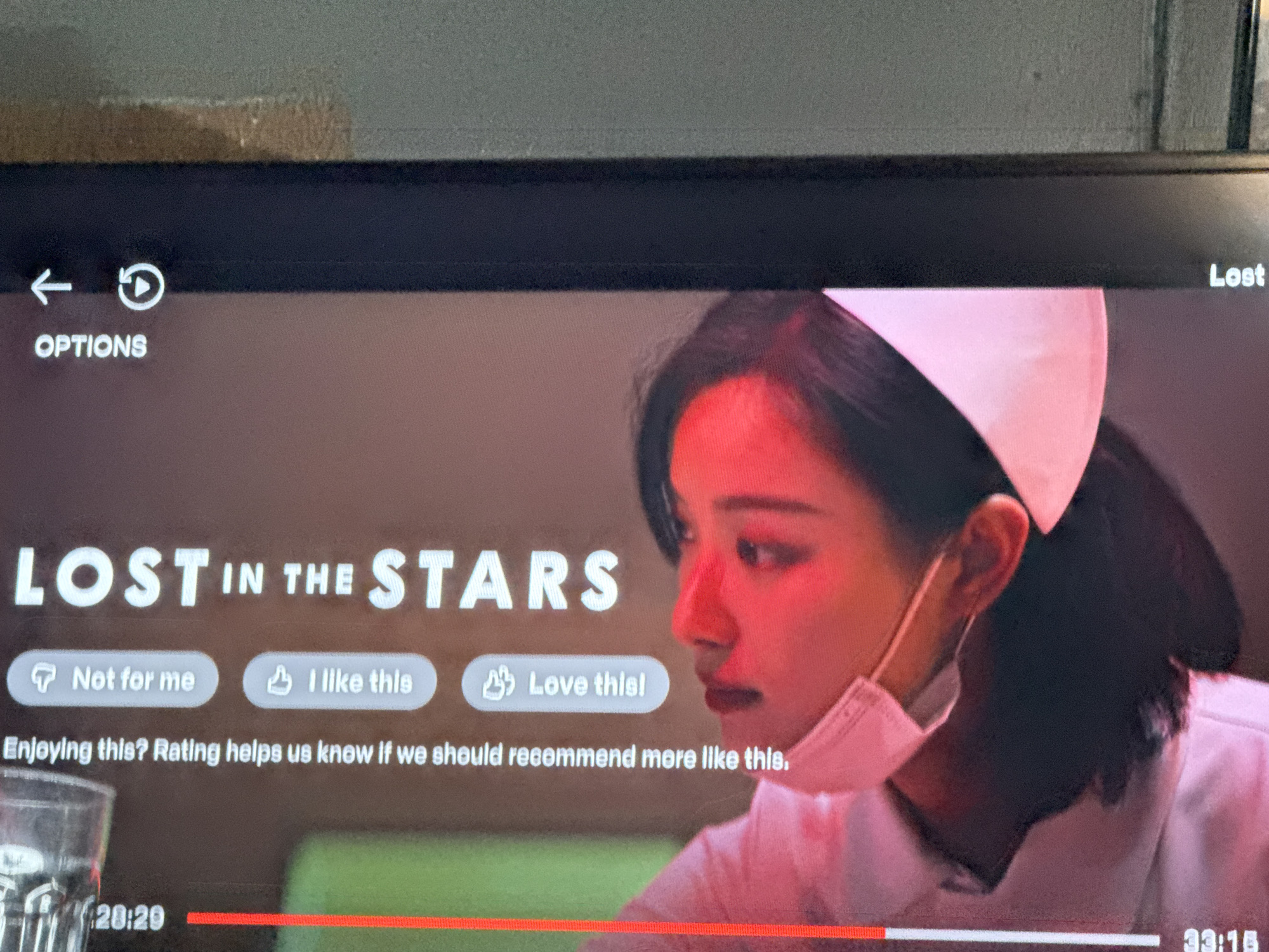The image captures a television screen displaying a paused scene from a movie or TV show, overlaid with a streaming service's user interface. The primary subject on the screen is a young East Asian woman, seen in profile from the chest up, wearing a white nurse-like uniform, a white cap, and a light pink surgical mask pulled down from her nose, exposing her mouth. Her face and hair are illuminated with a strong red light on one side, giving the scene a dramatic or stylized feel. Her expression appears serious or pensive. Her dark hair is pulled back, partially visible behind her ear.
The movie title, prominently displayed in large white letters, reads "LOST IN THE STARS", with "IN THE STA" slightly cut off but visible. Below the title, three interactive buttons offer rating options: "Not for me" with a thumbs down icon, "I like this" with a thumbs up icon, and "Love this!" with a heart icon. Below these, the text "Enjoying this? Rating helps us know if we should recommend more like this." is visible.
At the bottom of the screen, a red progress bar indicates the current viewing time at "20:20" and a total or end time of "33:16". In the top left corner, navigational elements include a left arrow, a refresh/play icon, and the word "OPTIONS". A fragment of the title "Lost" is visible in the top right.
A clear glass, partially filled with liquid, is visible in the bottom left foreground, suggesting the viewer is at home. The overall image depicts a moment of interaction with digital media, with the movie scene serving as the background for the interface. The external environment of Carmen, Philippines where this photo was taken is not depicted within the screen content itself.