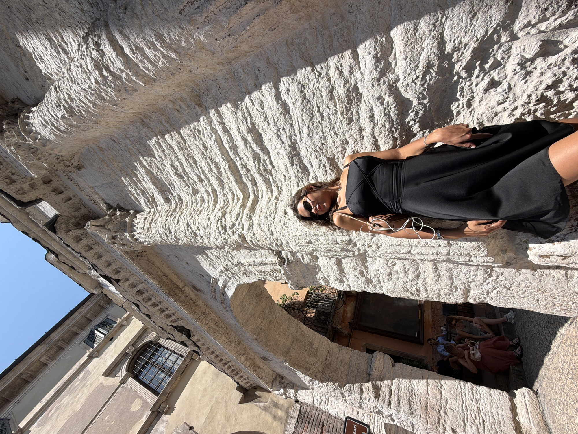 A woman in a black dress and sunglasses stands in front of a large, textured stone archway on a sunny day in Verona, Italy. The archway, made of rough-hewn, pale stone, is part of an ancient building with a tan facade and a barred window visible on the upper left. Underneath the archway, several people are seated in a recessed area. The woman is positioned on the right side of the frame, leaning against the stone. She is wearing a black sleeveless dress with crisscross straps on the bodice and carrying a light brown purse over her shoulder. White earbuds are visible around her neck. The scene is well-lit, with strong shadows cast by the architectural elements.