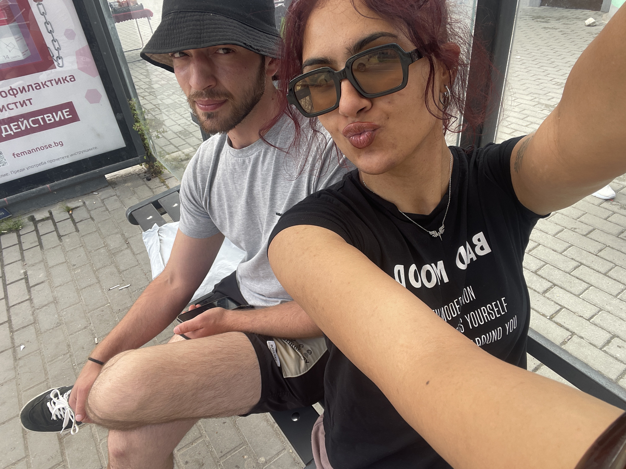 A selfie captures two individuals seated on a dark metal bench at an outdoor public location, likely a bus stop, in the city of Unknown, Unknown. The ground is paved with grey interlocking concrete pavers, showing some small debris.
The individual taking the selfie, a woman with auburn hair, is positioned in the foreground on the right. She wears large black-framed sunglasses with amber lenses, has dark eyebrows, and her lips are pursed in a kissing gesture. A silver hoop earring is visible in her left ear, and she wears a silver chain necklace with a small winged pendant. Her black t-shirt displays white text, partially legible as "OOD D", "OODED ON", "S YOURSELF", and "ROUND YOU". Her left bicep shows areas of lighter skin coloration and a faint tattoo. Her right arm is extended, holding the phone.
Seated to her left is a man with a short beard, wearing a black bucket hat, a grey t-shirt, and black shorts with a "BURBERRY" label visible on the left leg. He has a black bracelet on his right wrist and holds a black smartphone in his left hand. His legs are crossed, and he wears black sneakers with white laces. He is looking directly at the camera with a slight smile.
To the left of the man, a vertical advertisement board is visible. It features Cyrillic text in red on a white background, including "ПРОФИЛАКТИКА", "СТИСТИ", "ДЕЙСТВИЕ", and "femmanose.bg". Below, partially legible text reads, "Е. Преди употреба прочетете инструкциите." Small weeds grow at the base of the ad board. A single white shoe is visible on the far right edge of the paved ground.