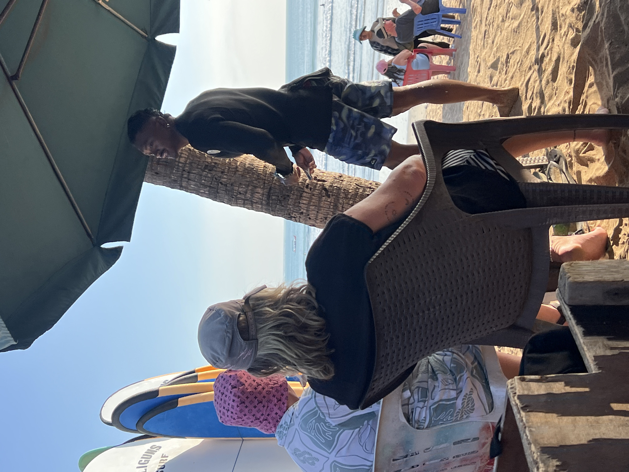 The image depicts a beach scene in Kuta, Indonesia. In the foreground, two individuals are seated facing away from the camera, their backs to the viewer. The person on the left wears a white patterned shirt and a pink bucket hat. The person on the right wears a black t-shirt and a grey cap, with blonde, wavy hair. They are seated in dark plastic chairs on a sandy beach.
Behind them, a man wearing a black long-sleeved top and blue patterned shorts stands next to a palm tree, looking to his right and holding a phone. The ocean and a clear blue sky are visible in the background, with small waves and distant boats. Further in the background, other people are seated on chairs, some on the sand. Surfboards are propped up on the left side of the image. An umbrella shades a portion of the scene. The lighting suggests it is daytime, likely late afternoon given the warm tones.