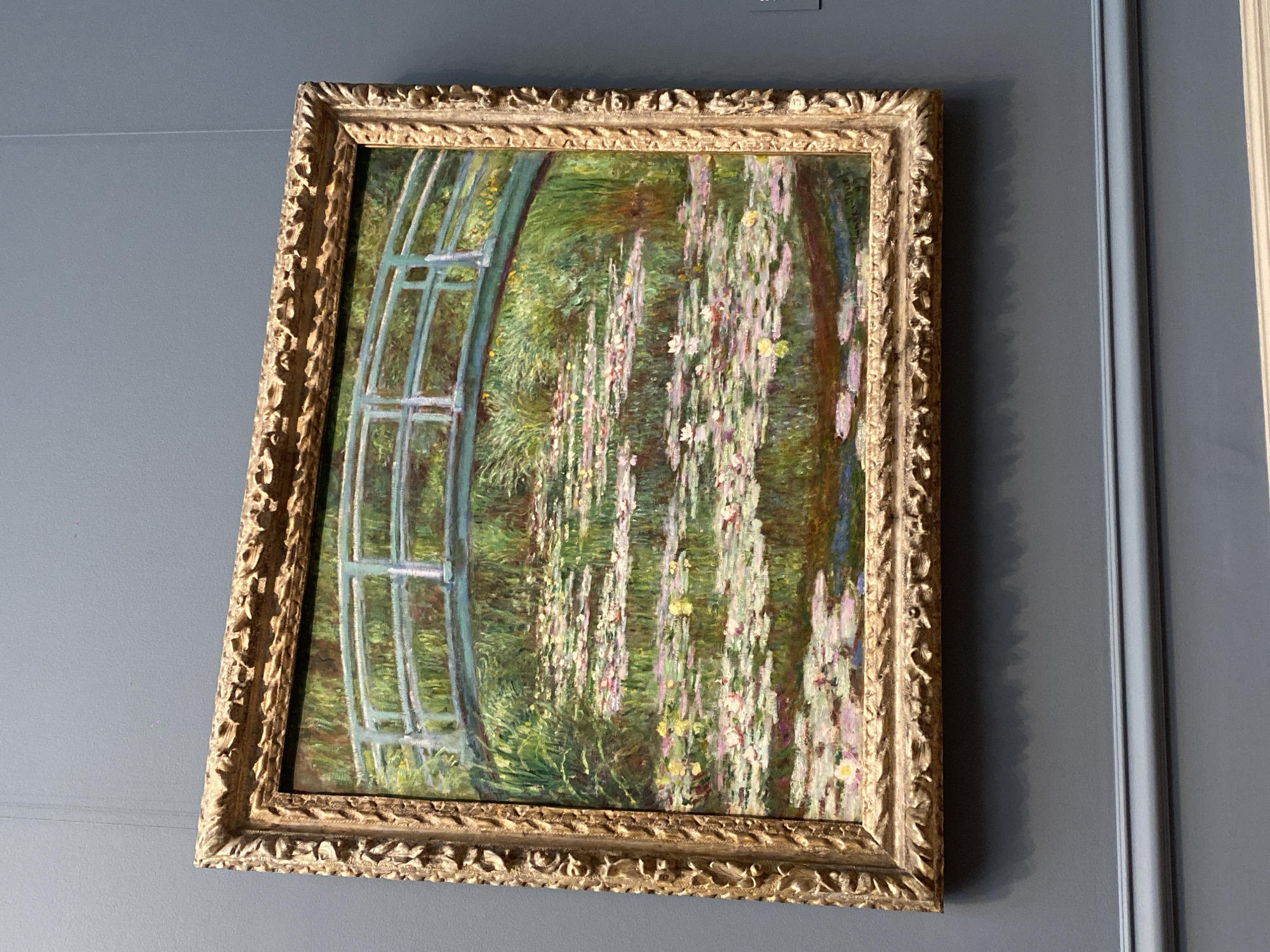 This is a photograph of Claude Monet's painting "The Water Lily Pond" housed in the National Gallery of Art in Washington, D.C. The painting depicts a serene pond filled with water lilies and a Japanese bridge arching over it, surrounded by lush greenery. The artwork is framed by an ornate, gilded frame and hangs on a muted gray wall. The lighting is ambient, and there are no people visible in the photograph.