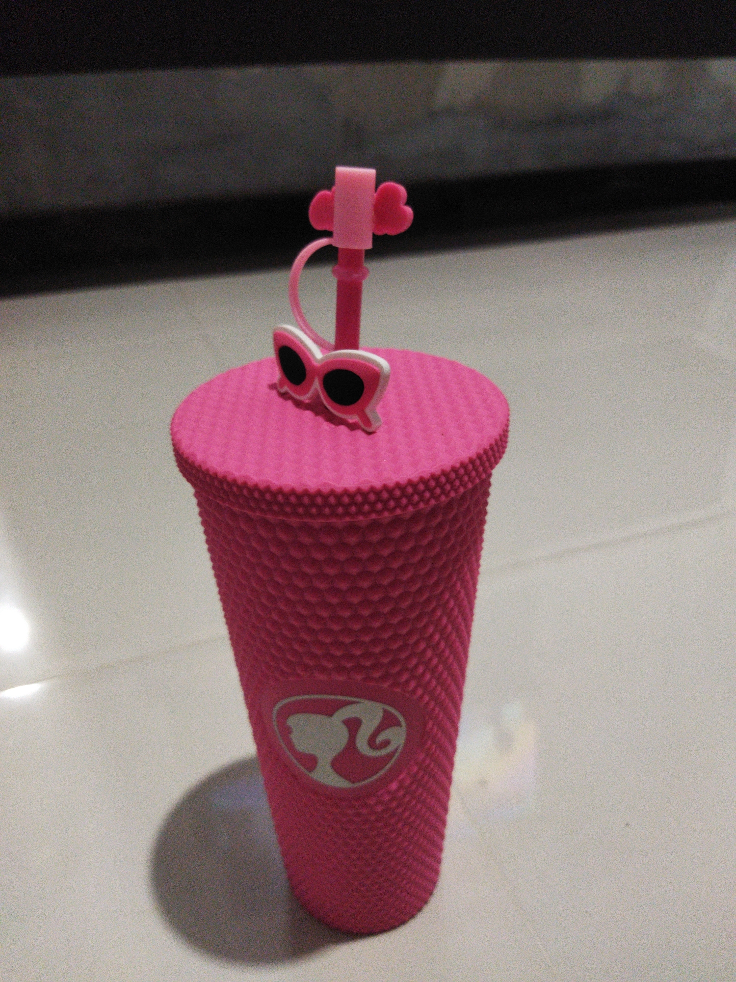 A pink Barbie-themed tumbler is placed on a light-colored, tiled floor. The tumbler has a textured, studded exterior and a pink lid with a straw. The lid is adorned with a small pink heart detail and a pair of white sunglasses with black lenses. A Barbie silhouette logo is visible on the lower portion of the tumbler. The background consists of a dark, out-of-focus area, possibly a wall or furniture. The image is taken from a slightly elevated angle, with a shadow of the tumbler cast on the floor to its left. The lighting appears to be artificial, creating a slight glare on the tiled surface. The specific location is not discernable from the image itself, and therefore its presence in Kosambi, Indonesia cannot be confirmed.