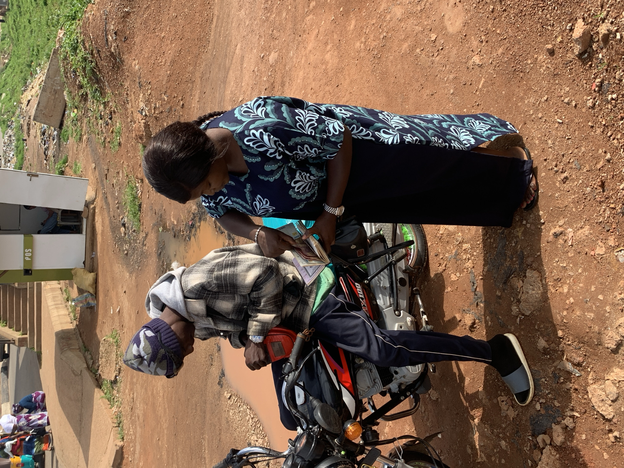 In Gura Top, Nigeria, a man wearing a camouflage beanie, plaid jacket, and dark pants with a white stripe sits on a motorcycle. He has a red pouch attached to the motorcycle's handlebars and appears to be reaching into it. A woman in a dark blue dress with a light blue floral pattern stands beside him, holding a book and what appears to be money. The ground is reddish dirt, with puddles of muddy water in the foreground. In the background, a building with a "POS" sign is visible, along with a set of stairs and scattered debris. The overall scene suggests an outdoor commercial or transactional interaction.