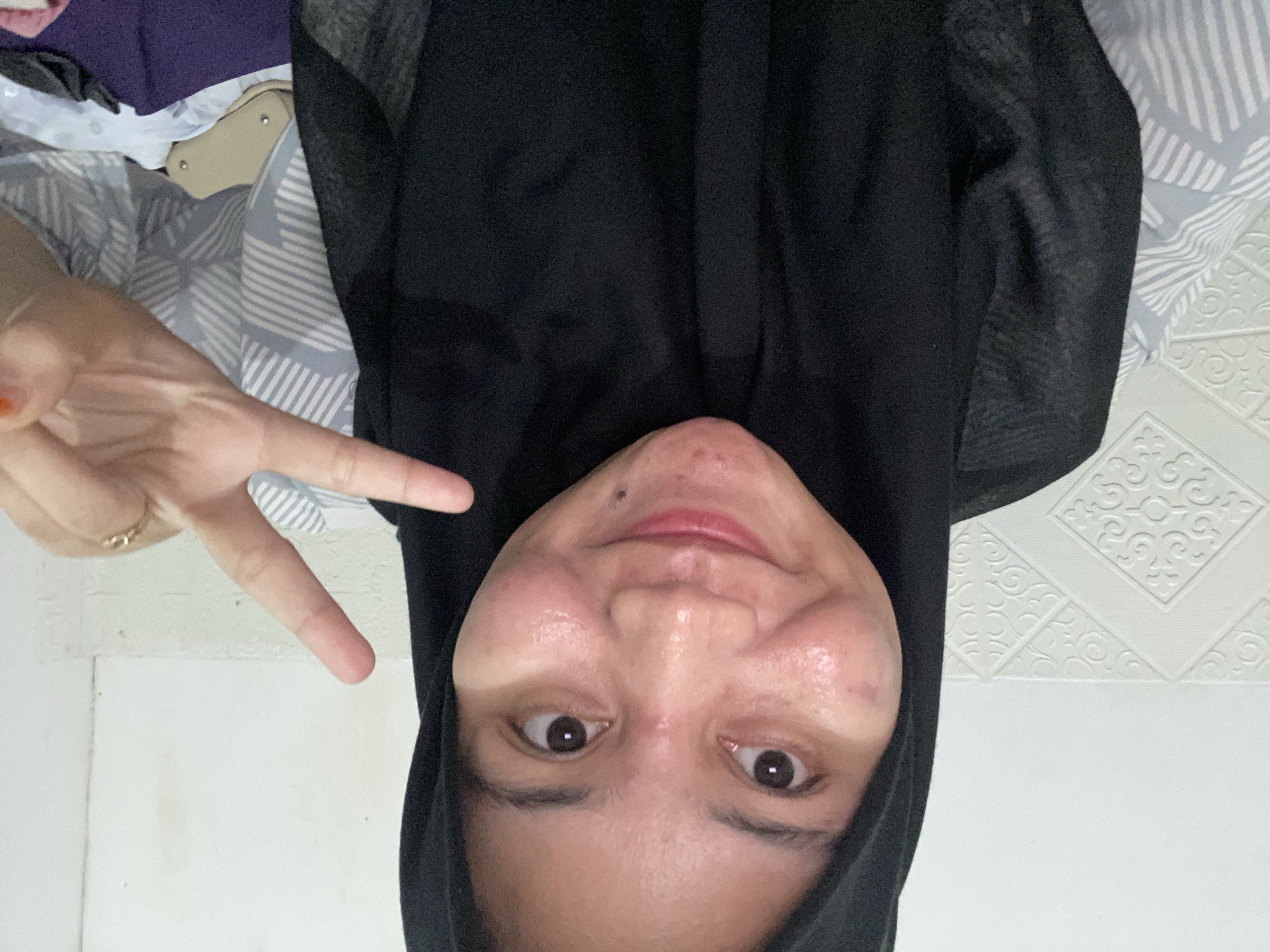 A young Indonesian woman wearing a black hijab and a black outer garment smiles at the camera. Her face is illuminated, revealing a fair complexion with some redness and blemishes on her cheeks and chin. She has dark eyes and is making a peace sign with her right hand, which is adorned with a delicate gold ring. The background shows a white textured wall and a patterned fabric. The lighting suggests an indoor setting, possibly a bedroom.