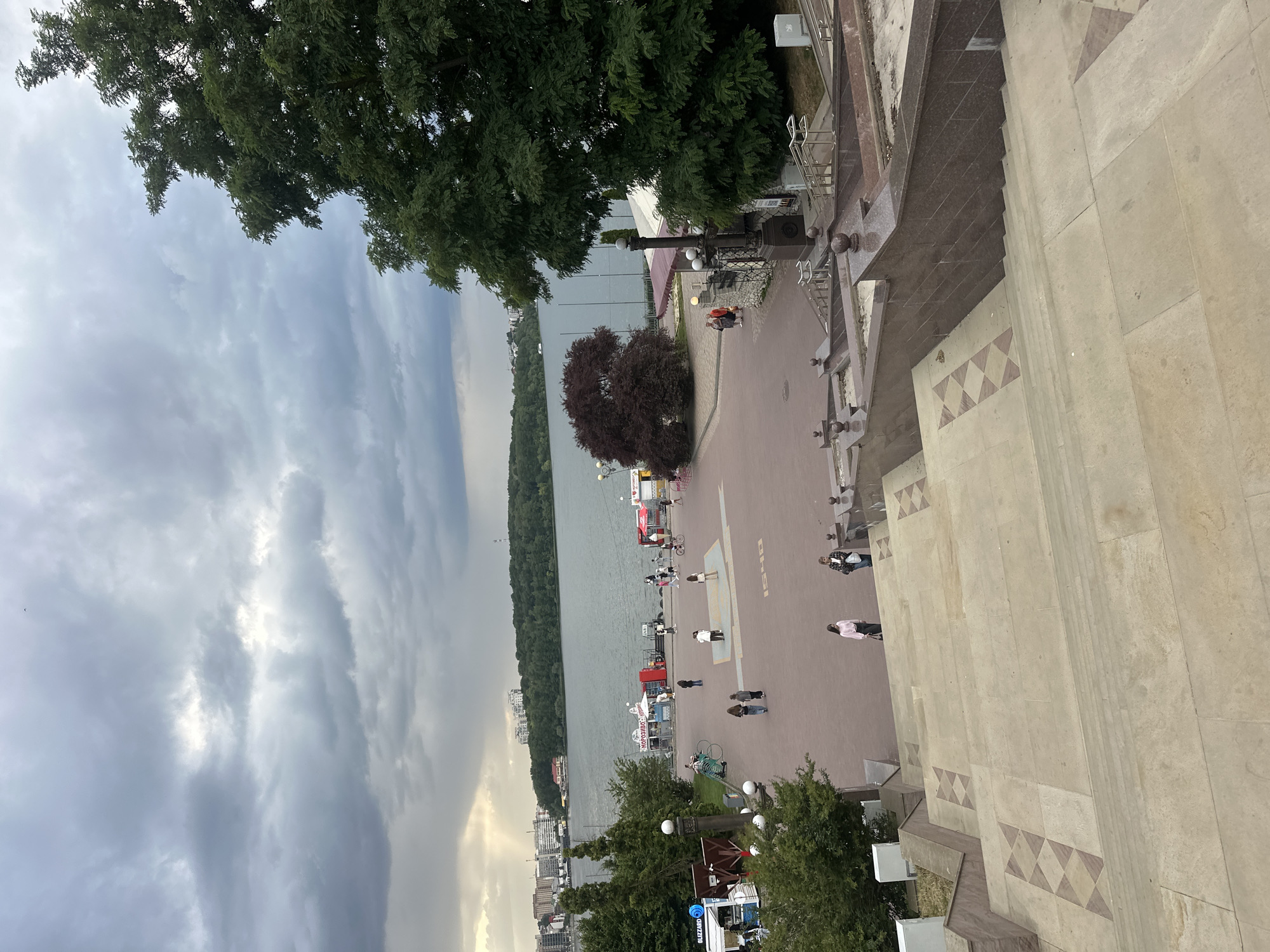 This is a vertical, medium shot of the Ternopil Pond embankment in Ternopil, Ukraine. The foreground is dominated by a wide, ornate stone staircase with patterned treads. The stairs descend towards a paved embankment area where several people are walking and standing. The embankment overlooks a large body of water, the Ternopil Pond, which fills the middle ground of the image. Across the pond, a tree-lined shore is visible, with buildings and a radio tower in the distance. The sky is overcast with dramatic, dark clouds, but a patch of sunlight breaks through in the upper left quadrant.
On the embankment, a woman in a white top and pink skirt walks towards the viewer. Further back, a group of people are gathered near a red kiosk. A bicycle is parked to the left of the kiosk. Another woman in a pink top and dark pants walks away from the viewer. Two other individuals are walking together further into the distance. There are also several food stalls and kiosks lining the embankment. A large, dark red tree stands prominently on the embankment, to the left of center. To the right, a large, leafy green tree partially obscures the view of the buildings and the pond.
The overall impression is one of a public recreational area on a potentially stormy day. The scene is captured from a high vantage point, looking down the staircase and across the pond.