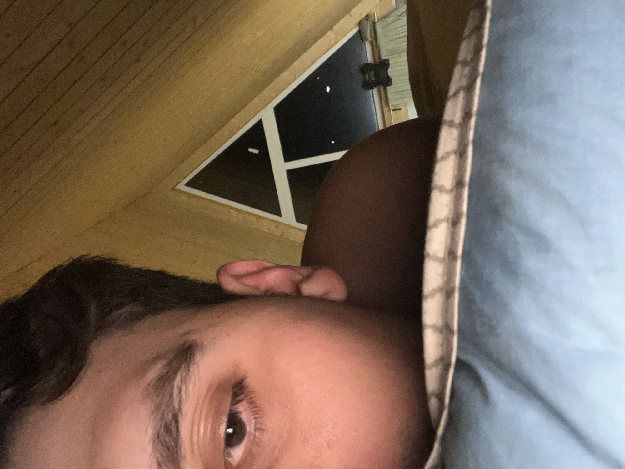 A close-up, low-angle, indoor shot reveals a person lying down in what appears to be an attic or room with a sloping wooden ceiling. A triangular window is visible, suggesting an unconventional architectural design. Only part of the person's face, including their eye, eyebrow, ear, and dark hair, is visible. They are resting on a pillow or cushion with a light blue cover with a textured edge. The lack of identifying information prevents confirmation of the location in Vasilyevo, Russia.
