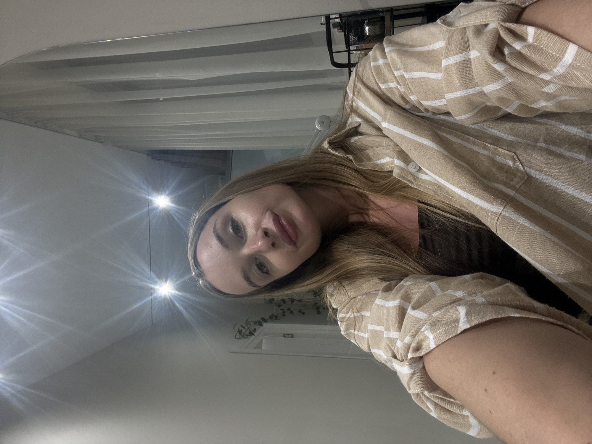 This selfie, rotated 90 degrees counter-clockwise, features a young woman with long, light brown hair, captured from the chest up. She looks directly towards the camera with a neutral to slightly smiling expression. She is wearing a casual, light beige or tan button-up shirt with prominent white horizontal stripes, and a darker garment is visible underneath. Her right arm, covered by the rolled-up sleeve of the striped shirt, is visible in the foreground.
The image appears to be a mirror selfie, with the background primarily consisting of reflections. Several bright light sources, likely indoor ceiling lights, are strongly reflected in the mirror, creating distinct starburst flare effects across the upper and left portions of the frame (if viewed upright). These reflections illuminate the indoor setting, which includes white horizontal blinds or shutters and glimpses of what appears to be a shelving unit or wire rack with objects, possibly in a living space or bedroom. A small plant is also faintly visible in the reflection. The scene is illuminated by artificial light, suggesting an indoor environment, potentially in Budva, Montenegro. No visible text is present in the image.