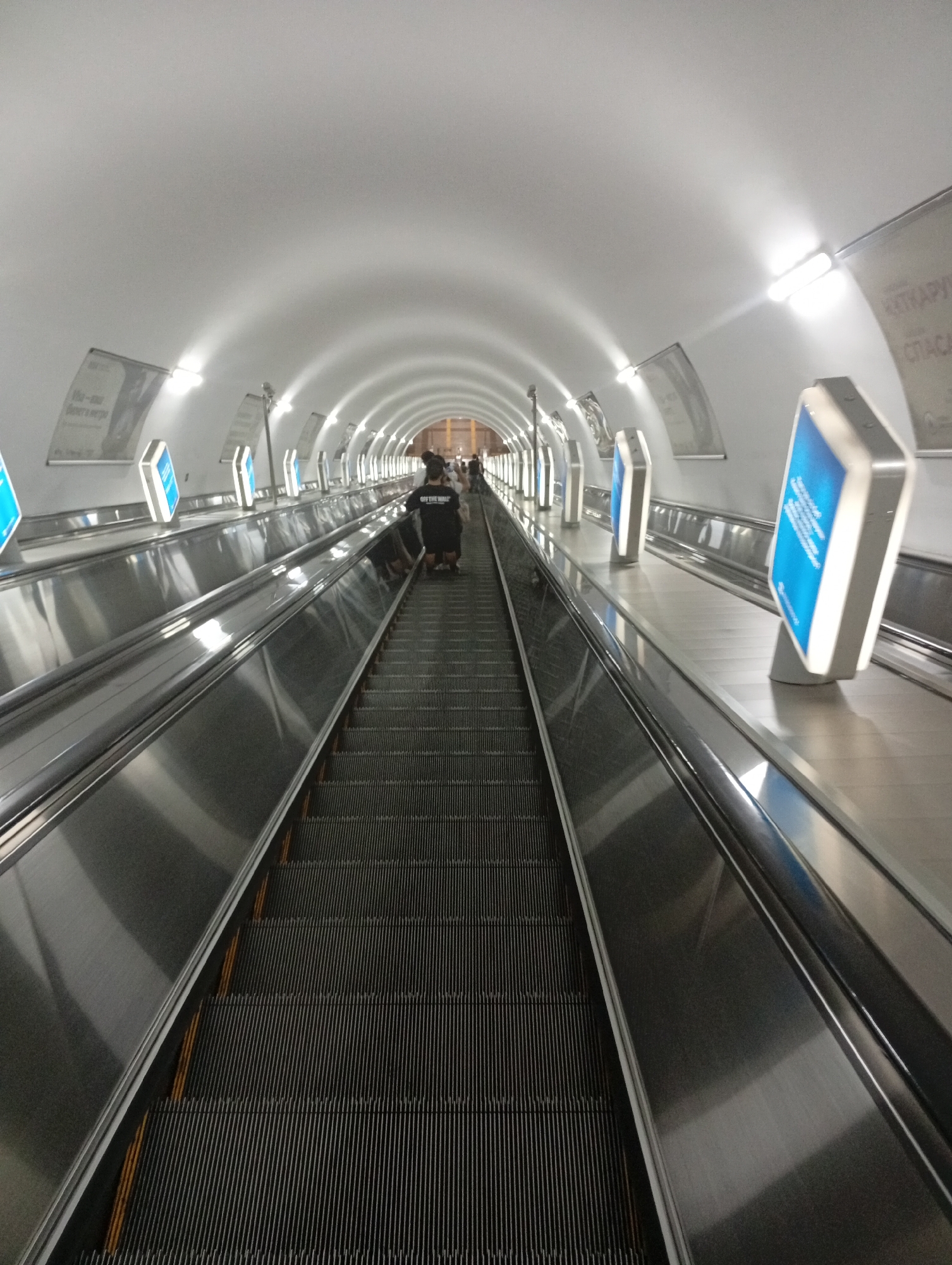 The image depicts a view looking down a long, brightly lit escalator in a subway station. The escalator is flanked by sleek, metallic handrails and advertisements. Two individuals, one wearing a black t-shirt with "OFF THE WALL" printed on the back, are visible on the escalator, appearing to be going down. The tunnel-like ceiling curves overhead, illuminated by numerous fluorescent lights, creating a futuristic and cavernous atmosphere. The escalators are on parallel tracks, suggesting a multi-track station. This is likely the escalator at the "Alatau" metro station in Almaty, Kazakhstan, known for its deep escalators and distinctive architecture.