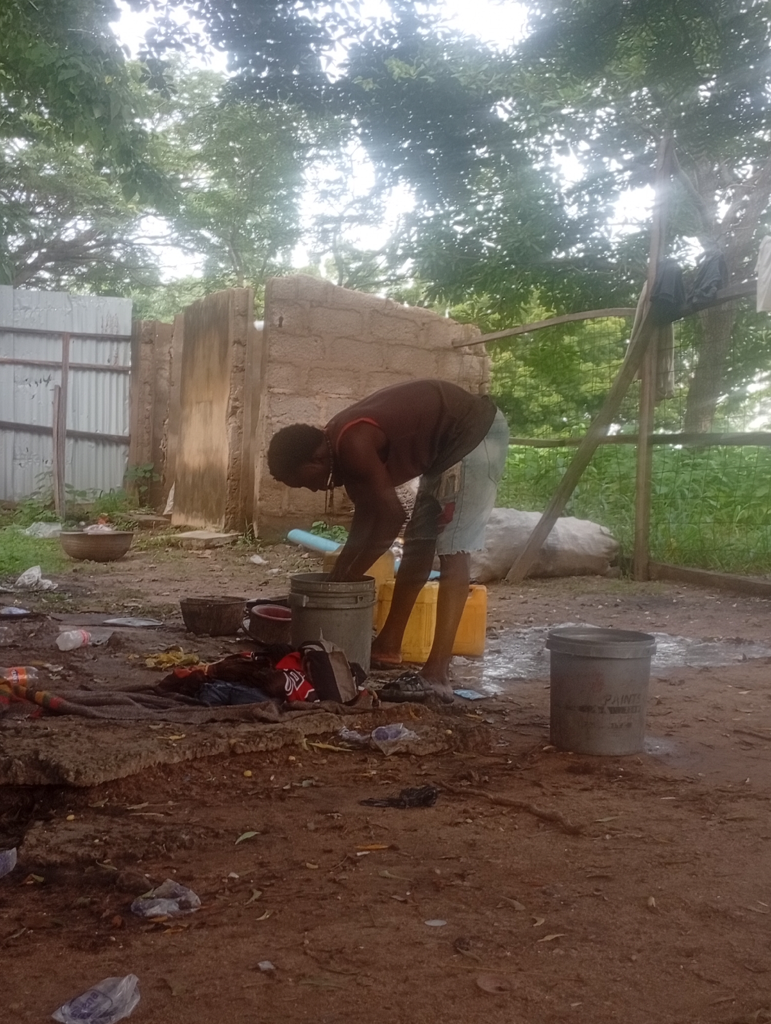 In Adankolo, Nigeria, a person with dark skin wearing a dark red tank top and light blue denim shorts is bent over, appearing to wash something in a gray metal bucket. The bucket sits on a dirty, uneven ground covered with loose soil, leaves, and scattered debris, including plastic bottles and wrappers. To the left of the bucket is a large, yellow plastic container. Further left, a shallow, wide brown bowl holds some items. In the foreground, a dark, rough blanket or fabric is spread out, with clothing items and a black backpack on top of it. Behind the person, a rough, reddish-brown mud-brick structure with an opening stands, and beyond that, a corrugated metal fence. The environment appears to be outdoors, with lush green trees and foliage visible in the background under a bright sky, suggesting an overcast day. A wooden frame structure, possibly part of a shelter, is also present to the right.
