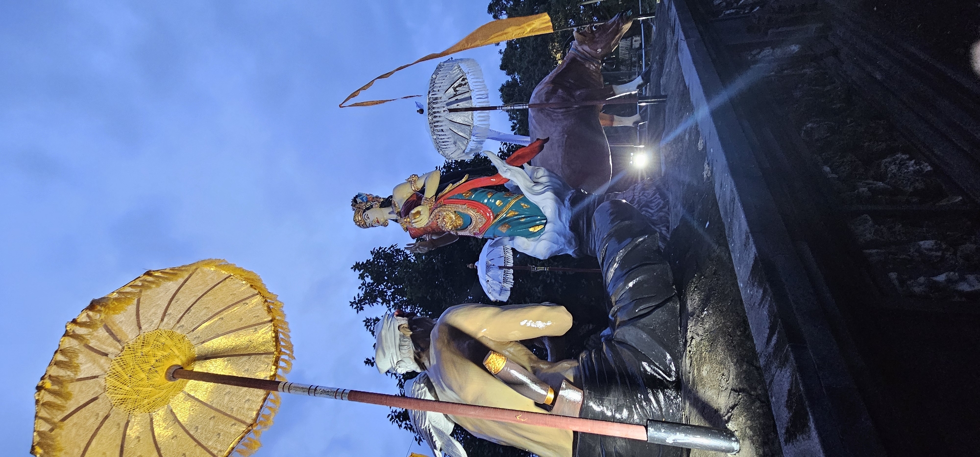 The image displays a vibrant outdoor scene featuring large, traditional Balinese statues under a clear, light blue sky, characteristic of either early morning or late afternoon. Artificial lights illuminate the statues and surrounding structures, suggesting it may be transitioning into evening.
In the foreground, a prominent golden ceremonial umbrella (payung) with fringed edges and intricate patterns is visible on the left. Adjacent to it stands a male statue, possibly a warrior or guardian, depicted from the side. He wears a white head covering, a light-colored upper garment, and dark lower attire, with a golden-hilted weapon at his waist.
The central focus is a graceful female deity or noblewoman statue, elaborately adorned with a crown or intricate headdress, golden jewelry, and richly patterned clothing in blue-green, red, and gold. Her hands are positioned in front, possibly in a gesture of offering or blessing. Behind her, a large brown animal statue, resembling a bull or cow with white legs, faces upwards.
Several other white ceremonial umbrellas are scattered around the statues, and a yellow flag or banner is seen fluttering in the background. Dark green foliage from trees provides a natural backdrop. The statues are situated on a textured stone or concrete platform that forms a wall-like structure on the right, suggesting a temple complex or public display area. The overall scene evokes a sense of cultural and religious significance, typical of Denpasar, Indonesia. No visible text is present.