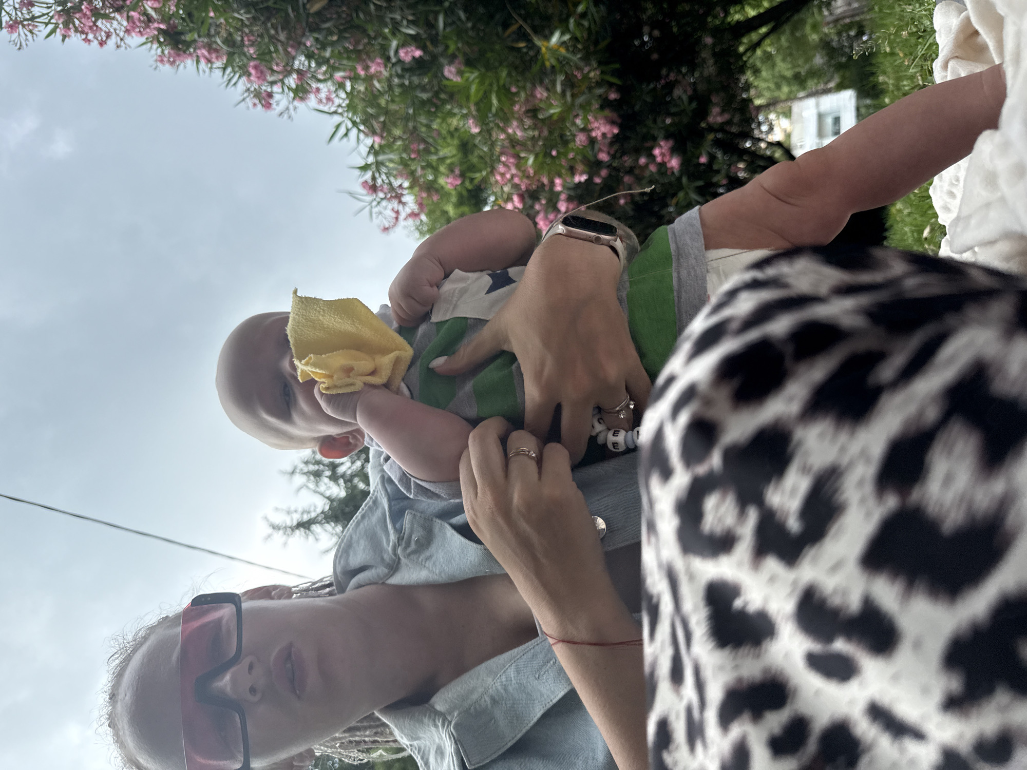 The image shows a close-up view of two people, an adult and a baby, in an outdoor setting. The adult, wearing sunglasses and a light blue shirt, is holding the baby. The baby is wearing a green and white striped shirt and has a yellow cloth toy. The background features greenery and pink flowers, suggesting a garden or park. The sky is overcast. The perspective is from below, looking up at the subjects. The image appears to be taken during the daytime. The presence of a red string bracelet on the adult's wrist might be a cultural or symbolic detail. The overall mood seems casual and familial.