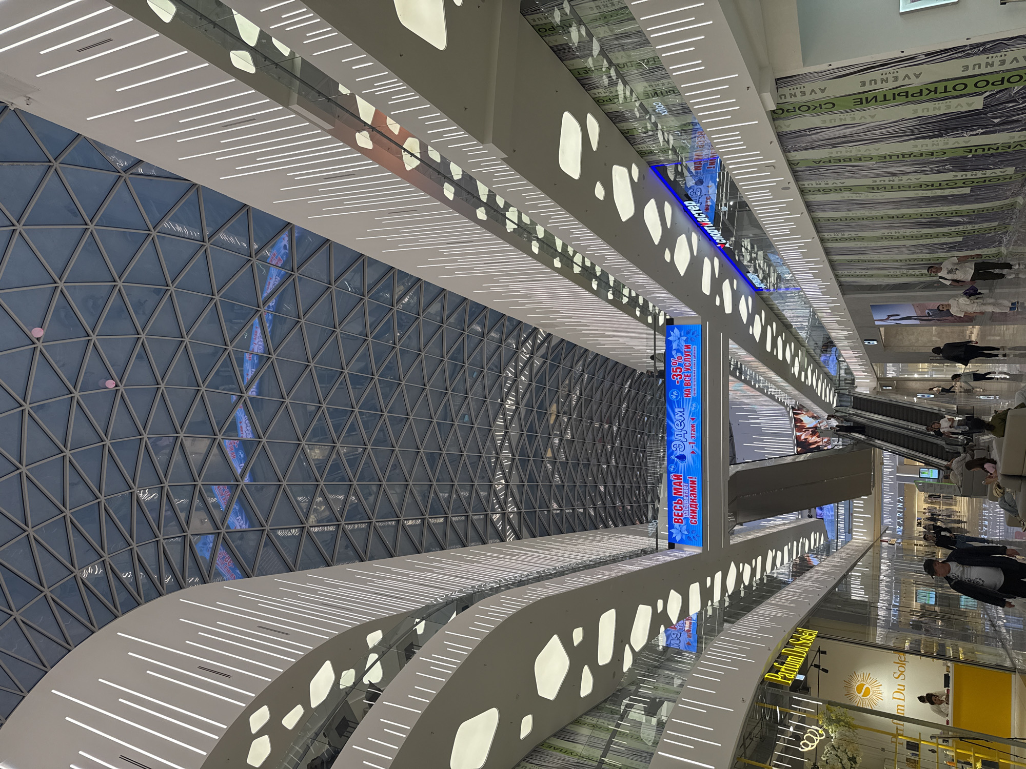 This is an interior shot, likely within a shopping mall or atrium, possibly located in Moscow, Russia based on Cyrillic text visible on signs. The architectural design features a geodesic dome structure to the left, constructed from triangular panes. The interior spaces are multi-tiered with balustrades exhibiting linear and organic-shaped light fixtures.
Advertisements and signage are interspersed throughout, with promotions written in Russian. Escalators are visible, suggesting movement between floors. The mall seems active, with several people walking and shopping on different levels. Several shops and retail spaces can be identified, including a "Parfum Du Soleil" storefront.