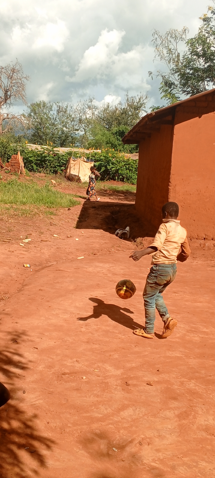 An outdoor scene in Ngalula, Democratic Republic of the Congo, depicts a sunlit, unpaved area of reddish-brown dirt. In the foreground, a child, viewed from behind, wears a light-colored short-sleeved shirt, dirty blue jeans, and light-colored sandals, engaged in kicking a multi-colored (black and brown/gold) soccer ball with their right foot. A distinct shadow of the child and the ball is cast on the ground.
To the right, a building with reddish-orange earthen walls and a dark roof is partially visible. Near its base, a dark-colored object resembling a discarded helmet or bag rests on the ground. Scattered across the dirt ground in the mid-left are several light-colored elongated objects, possibly corn cobs, and a crumpled metallic item.
In the background, a person, likely an adult, wearing a patterned dress, walks from left to right. Behind them and to their left, a dark-colored animal, consistent with a pig, is visible near a crude, light-colored, tent-like structure. Further back, dense green vegetation, including trees and shrubs, forms a natural backdrop under a sky with prominent white clouds and patches of blue.
