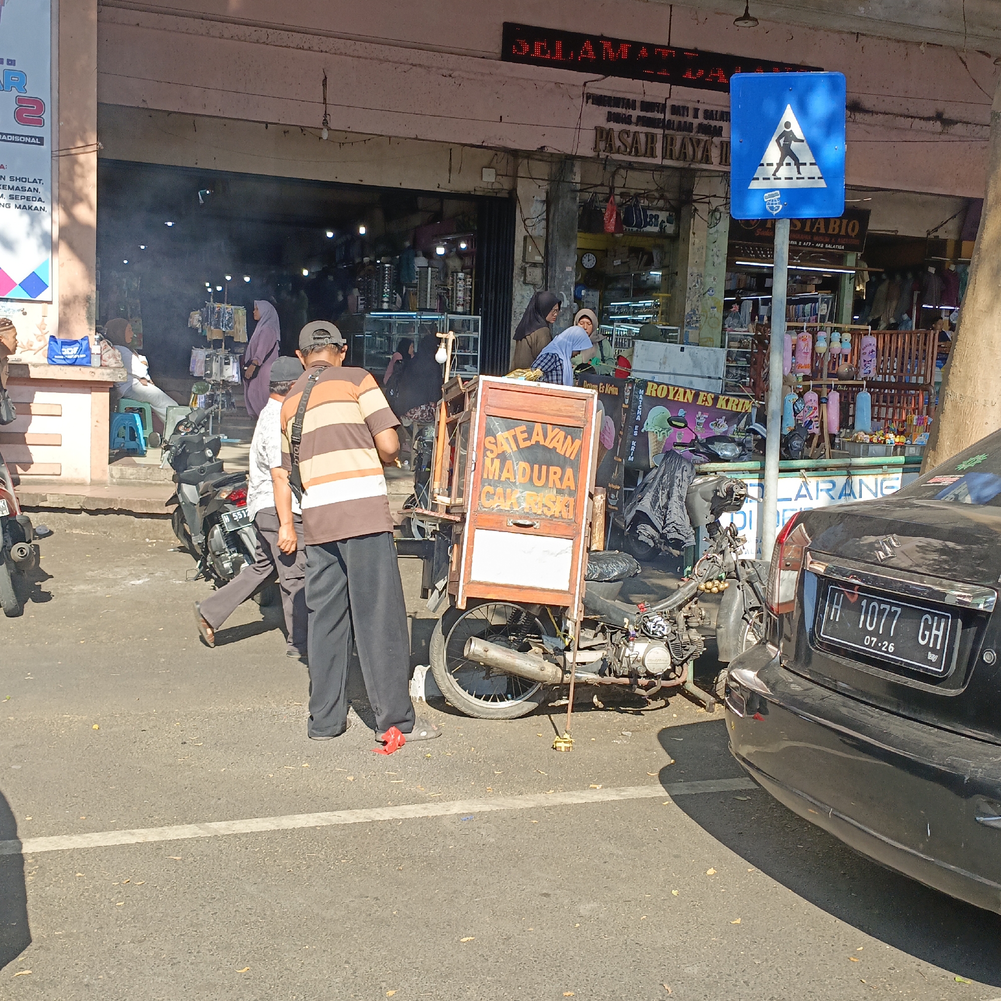 The image captures a street scene in Salatiga, Indonesia, likely in front of a market or shop. A street vendor's cart, advertising "SATE AYAM MADURA CAK RISKI," is prominently displayed, mounted on a motorcycle. The cart is made of wood and has signs with text and images of food. Several people are visible, including a vendor attending to the cart and customers browsing nearby. Other motorcycles and a black car with license plate "H 1077 GH" are parked on the street. In the background, shops with various goods are open, and a blue pedestrian crossing sign is visible. The environment appears to be a busy urban area with the presence of commercial activity.