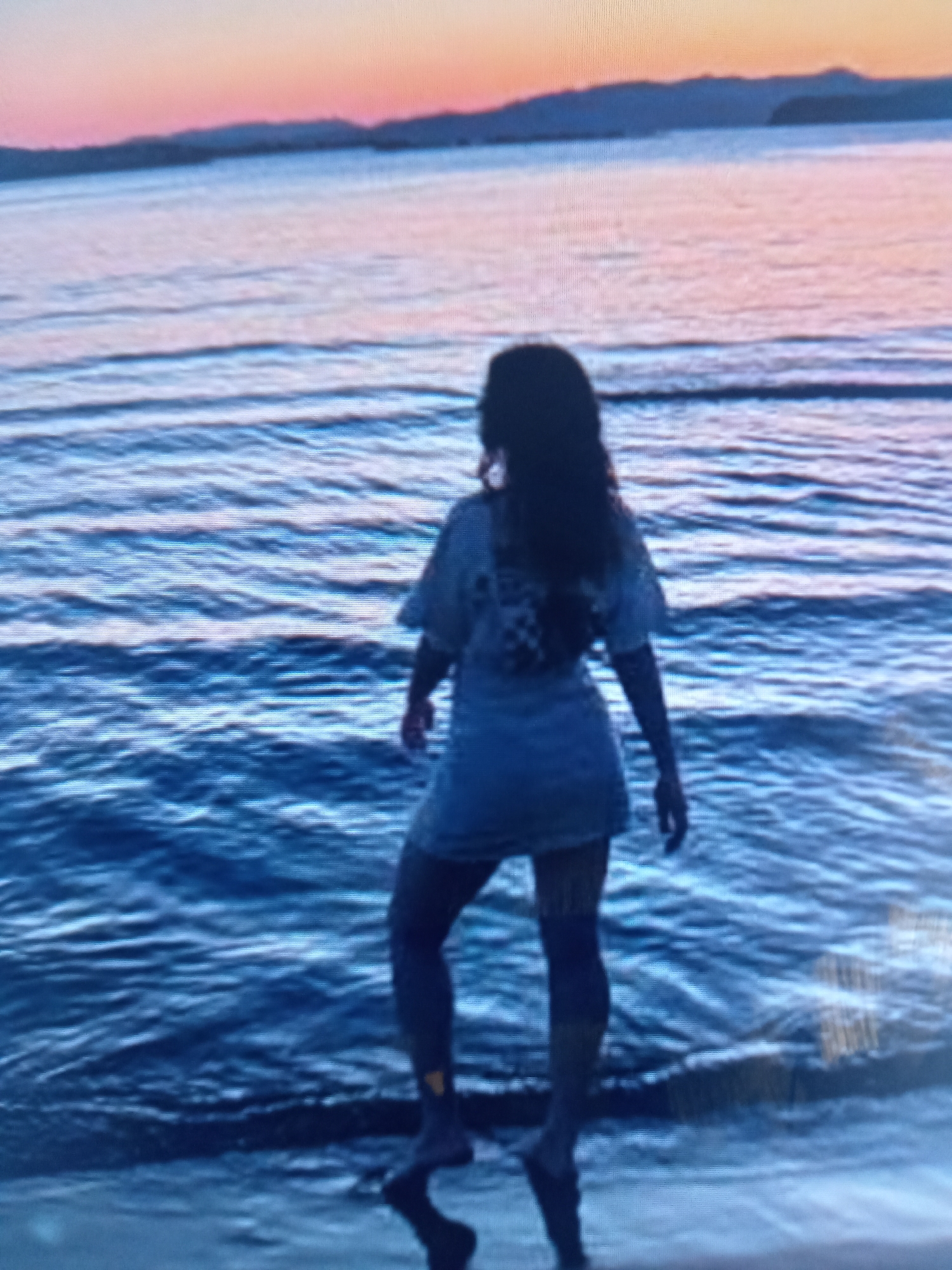 A person stands at the edge of the water, facing away from the camera, towards a sunset over the ocean. The person appears to be a woman with long, dark hair, wearing a light-colored t-shirt dress. Her legs are visible, and she is standing with her feet in the shallow water. The ocean is a deep blue, with small waves breaking on the shore. The sky is a gradient of pink, orange, and purple, with a faint silhouette of mountains in the distance. The overall mood of the image is peaceful and serene.