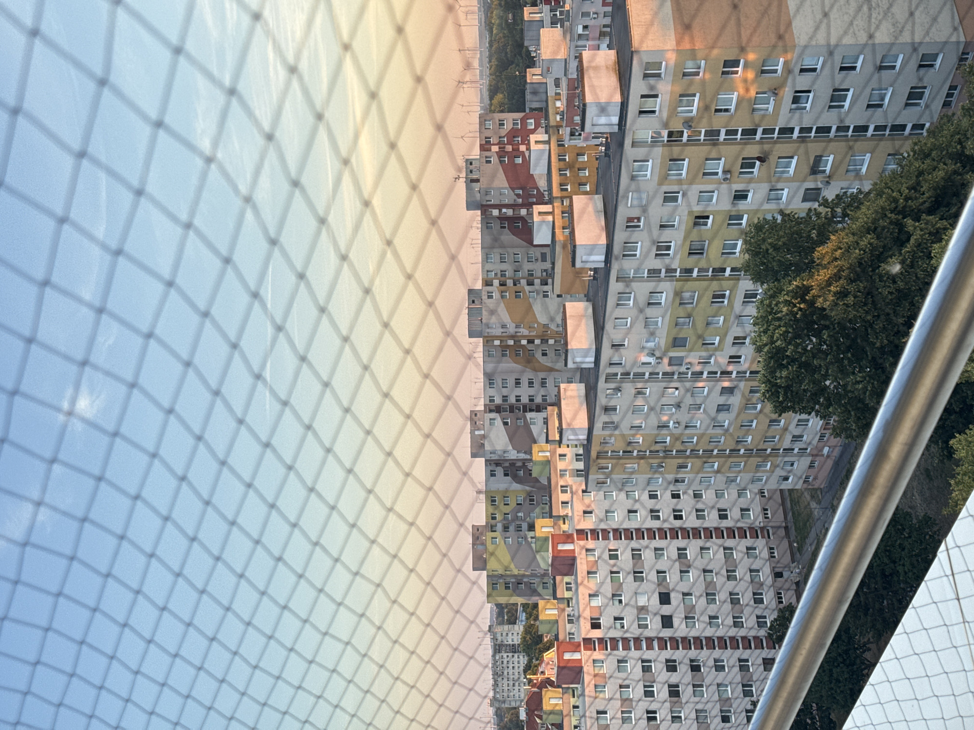 This image provides an elevated view of a residential area, likely in Bratislava, Slovakia, as indicated by the characteristic socialist-era apartment blocks. A meshed safety net in the foreground, likely from a balcony or rooftop, slightly obscures the scene. The apartment buildings are tall and multi-storied, painted in a variety of pastel colors, including yellow, pink, and grey, with some featuring geometric patterns in contrasting colors like red and brown. The buildings are densely packed, creating a cityscape. In the background, a line of wind turbines is visible on the horizon under a sky that transitions from pale blue at the top to warm hues of orange and pink at the bottom, suggesting either sunrise or sunset. Trees are visible in the foreground and between some of the buildings, indicating the presence of green spaces within the urban environment. There are no visible people or specific actions taking place. The overall scene depicts a residential urban landscape during a transitional time of day, characterized by a blend of modern architecture and natural elements.