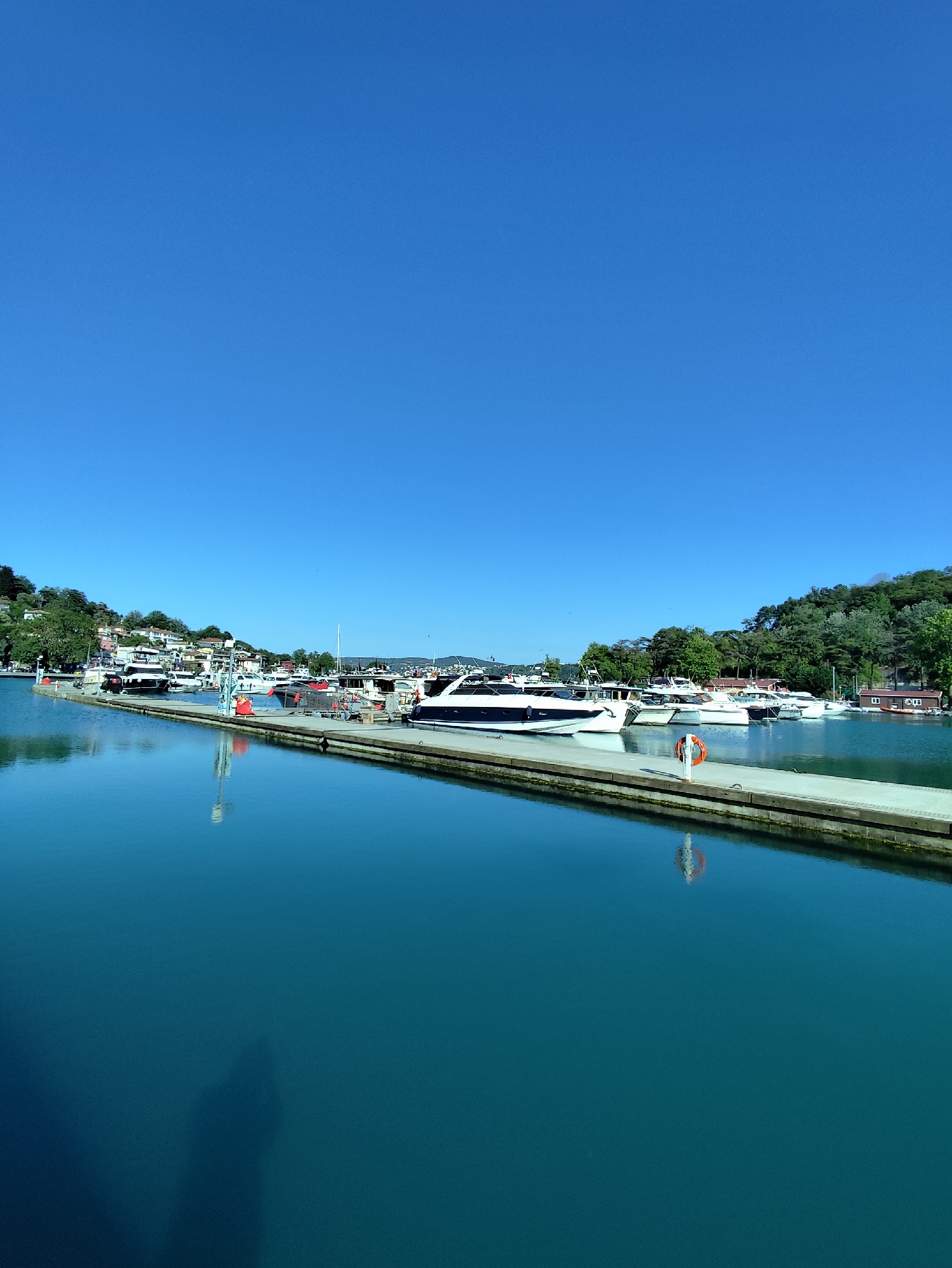 The image displays a marina in Sarıyer, Turkey, under a clear, deep blue sky. A light-colored concrete pier extends diagonally from the lower-left foreground towards the mid-right, separating the calm, blue-green water in the immediate foreground from the main docking area. Numerous white motor yachts and smaller cruisers are moored along the pier and further into the harbor. On the left and central background, a green, tree-covered hillside ascends, featuring several multi-story buildings with light facades and red or terracotta roofs nestled among the foliage. The right background consists of a densely forested hillside, with a small, single-story light-colored wooden structure with a red roof visible at the water's edge. A white lifebuoy with an orange ring is attached to a post on the pier, and its reflection is clearly visible in the tranquil water. The shadow of an individual, presumably the photographer, is cast upon the water in the lower-left quadrant of the image. No other individuals are overtly visible on the pier, boats, or shorelines.