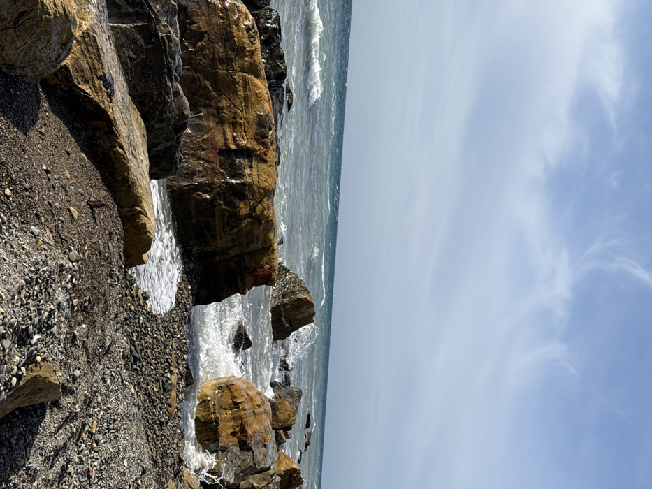The image presents a detailed view of a rocky coastline in Anapa, Russia, under a bright, partly cloudy sky. The horizon delineates a turbulent, grey-blue sea with discernible wave formations. Numerous large boulders, predominantly dark brown, black, and reddish-orange with visible striations, occupy the foreground and midground. These rocks are wet, indicating recent contact with the sea. White foam from actively breaking waves is visible around and between the boulders, particularly in the midground where water splashes against them. The immediate shoreline consists of a dark grey to black pebble and gravel beach, transitioning into larger rock formations at the water's edge. No human subjects or man-made structures are present within the frame. The scene is illuminated by natural daylight.