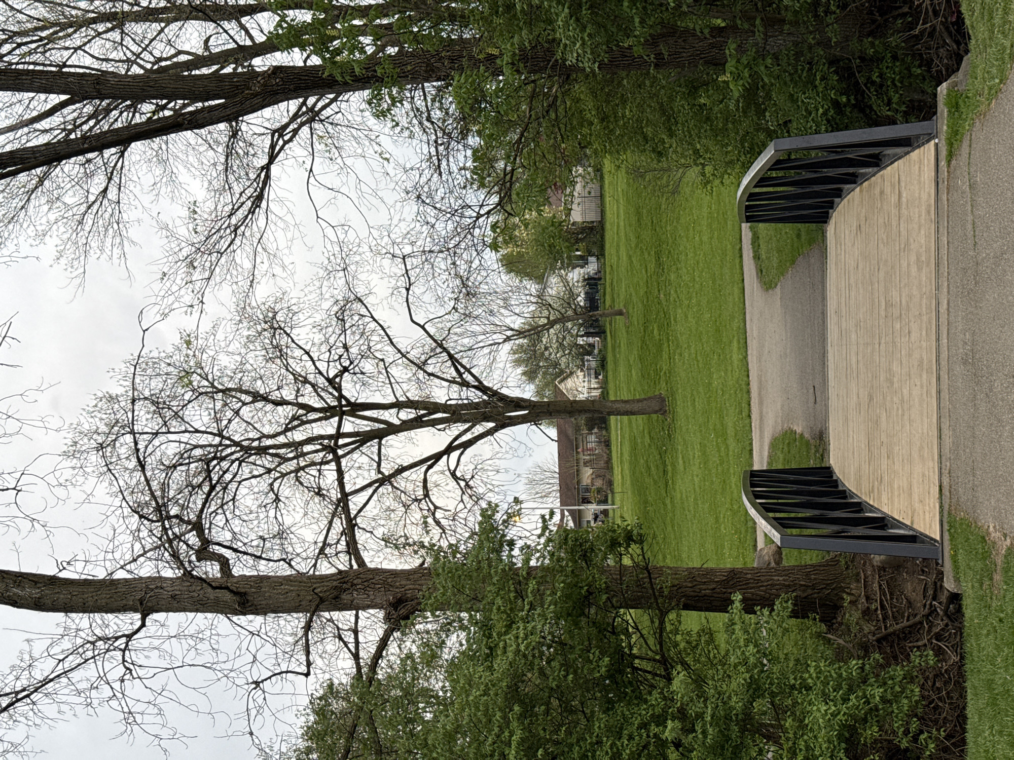 The image depicts a park scene, with a bridge spanning a green grassy area. The bridge has a wooden deck and dark metal railings. A paved pathway leads onto and off the bridge. Bare trees are visible on either side of the path, some with sparse new foliage. In the background, houses are seen across the lawn. The location is Grove City, Ohio, USA.