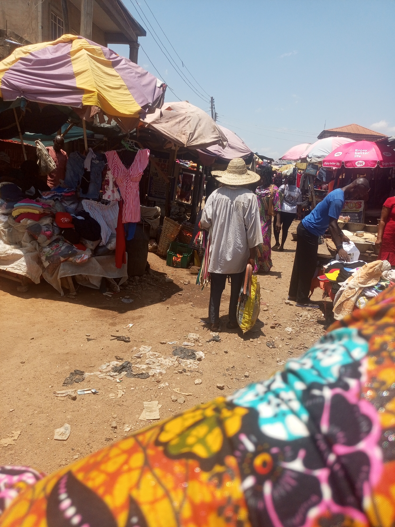 Here's a detailed description of the image:
**Setting:** The image captures an outdoor marketplace scene in Sabon Tasha, Nigeria, characterized by dirt and debris-strewn ground.
**People:**
* Several individuals are present, including shoppers and vendors.
* One person wearing a straw hat stands with their back to the camera, holding a shopping bag.
* Another individual in a blue shirt bends over a display of goods.
**Objects:**
* Numerous market stalls covered with various colored umbrellas.
* Merchandise on display includes clothing, fabric, and other goods.
* Bags, baskets, and containers are scattered throughout the scene.
* "Enjoy better life" branded umbrellas are visible.
**Actions/Interactions:**
* People appear to be browsing and shopping.
* Vendors are tending to their stalls.
**Environment:**
* The ground is dusty and littered.
* Buildings are visible in the background.
* Overhead power lines add to the scene's busy atmosphere.
* A section of patterned fabric is prominent in the foreground, partially obstructing the view.