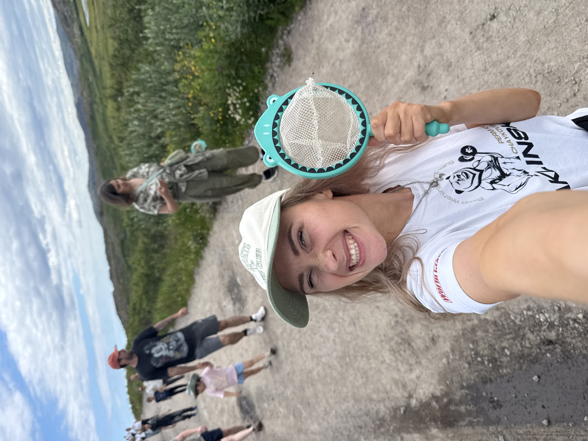 The image shows a group of people walking on a dirt road in Teriberka, Russia. In the foreground, a young woman is taking a selfie. She is wearing a white t-shirt and a baseball cap and holding a butterfly net. Behind her, other people are walking, including a man in a red cap and a child in a pink shirt. The landscape is characterized by green vegetation and rolling hills under a cloudy sky.