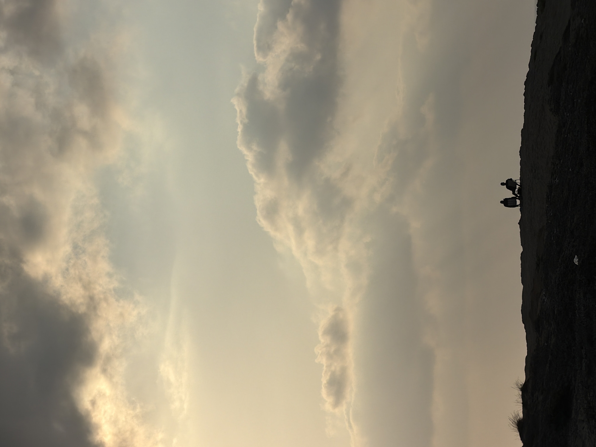 The image shows two silhouetted figures sitting on chairs on a dark, uneven terrain against a dramatic sky filled with clouds. The sky transitions from a pale yellow near the horizon to a darker grey towards the top. Sunlight filters through the clouds, creating a diffused, ethereal glow. The figures are positioned relatively close to each other on the crest of the hill, facing away from the camera towards the expansive sky. The terrain they are on appears barren and rugged, with some sparse vegetation visible on the left side. The overall mood is serene and contemplative, with the vastness of the sky contrasting with the intimate setting of the two individuals. The location is believed to be within the city of Hadera, Israel, likely a coastal or elevated area offering a wide view of the sky.