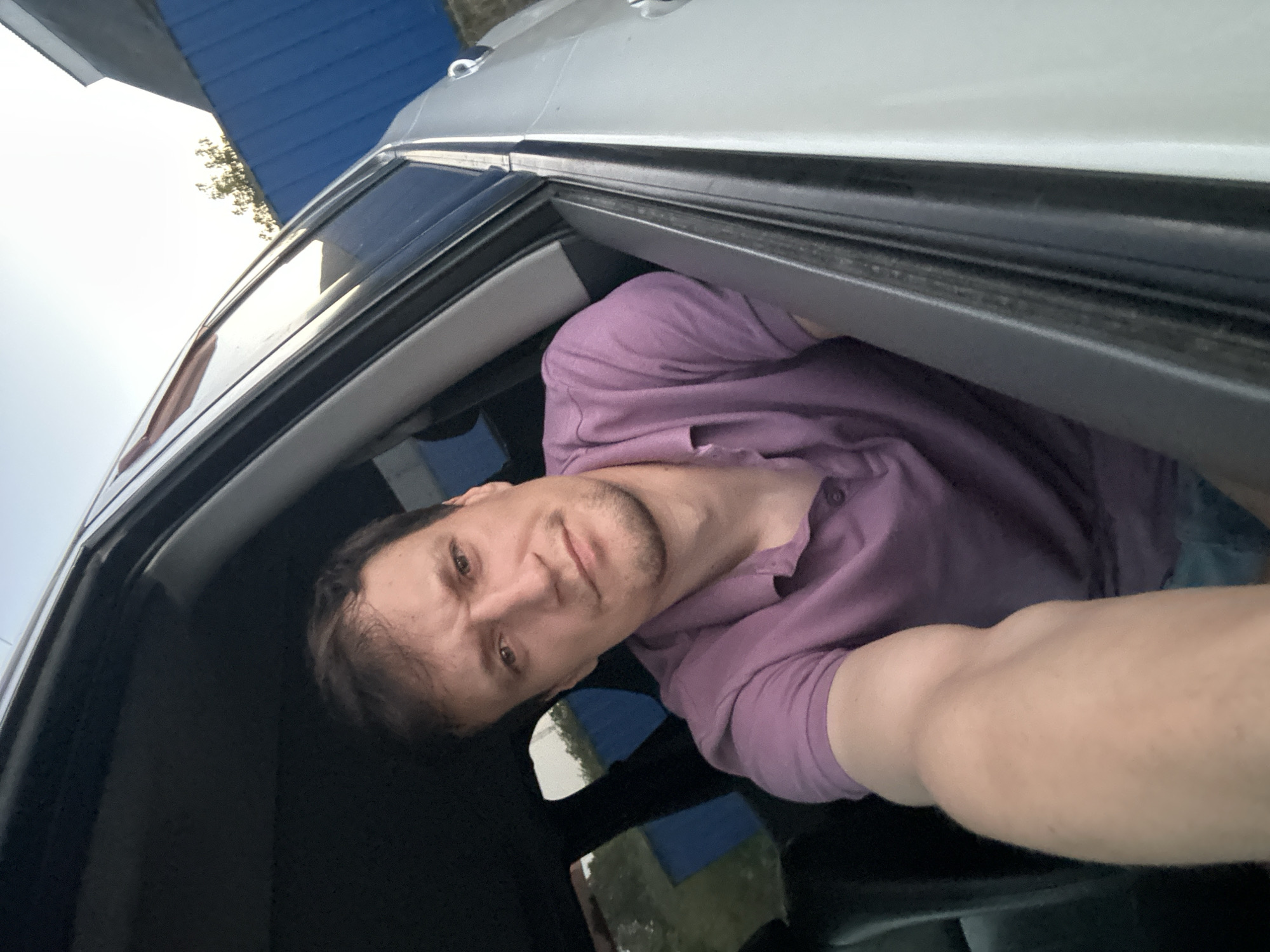 A man is seen from the chest up, peering out the open window of a car. He has short, dark hair and is wearing a purple polo shirt. His left arm is extended outwards, taking the photograph. The interior of the car is dark, with black seats visible. Outside the car, a blue fence and some greenery can be seen against a bright sky. The image appears to have been taken in Shatava, Ukraine.