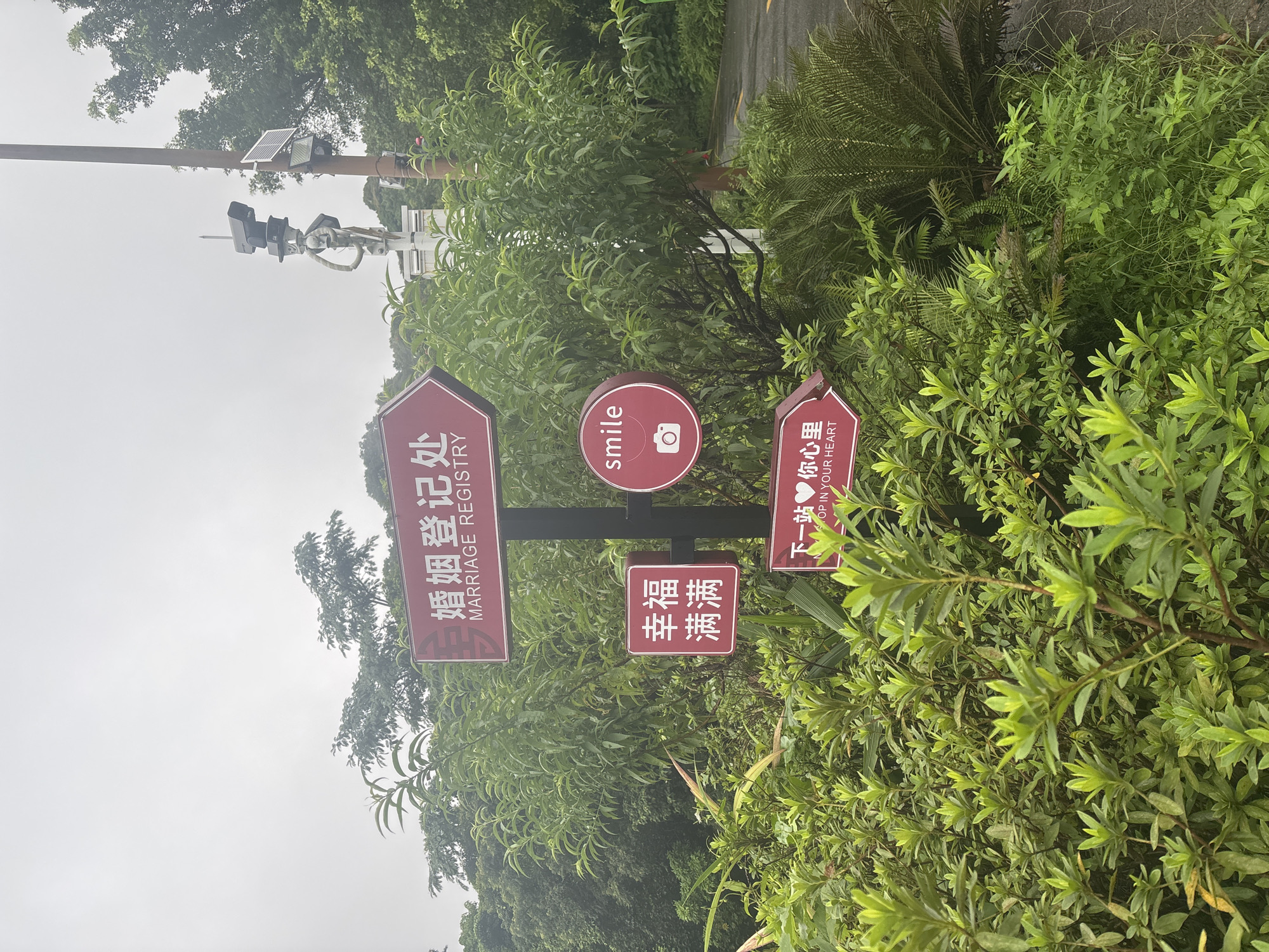 The image captures a cloudy day with lush green foliage in what appears to be a park or outdoor venue. Several red directional signs are prominently displayed on a metal pole. The topmost sign, shaped like an arrow, reads "MARRIAGE REGISTRY" in English and "婚姻登记处" in Chinese, indicating a place to register marriages. Below it, another red sign is square with the characters "幸福满满" which translates to "Full of Happiness". To the right of that, a circular red sign features the word "smile" in English and a camera icon, suggesting a photo opportunity. A final red arrow-shaped sign, positioned lower, says "下一站♥你心里" which translates to "Next stop ♥ in your heart". The background shows trees and a pathway, and a utility pole with multiple cameras and a solar panel is visible on the right. The overall impression is a scenic and possibly romantic location. This scene is located in Guangzhou City, China.