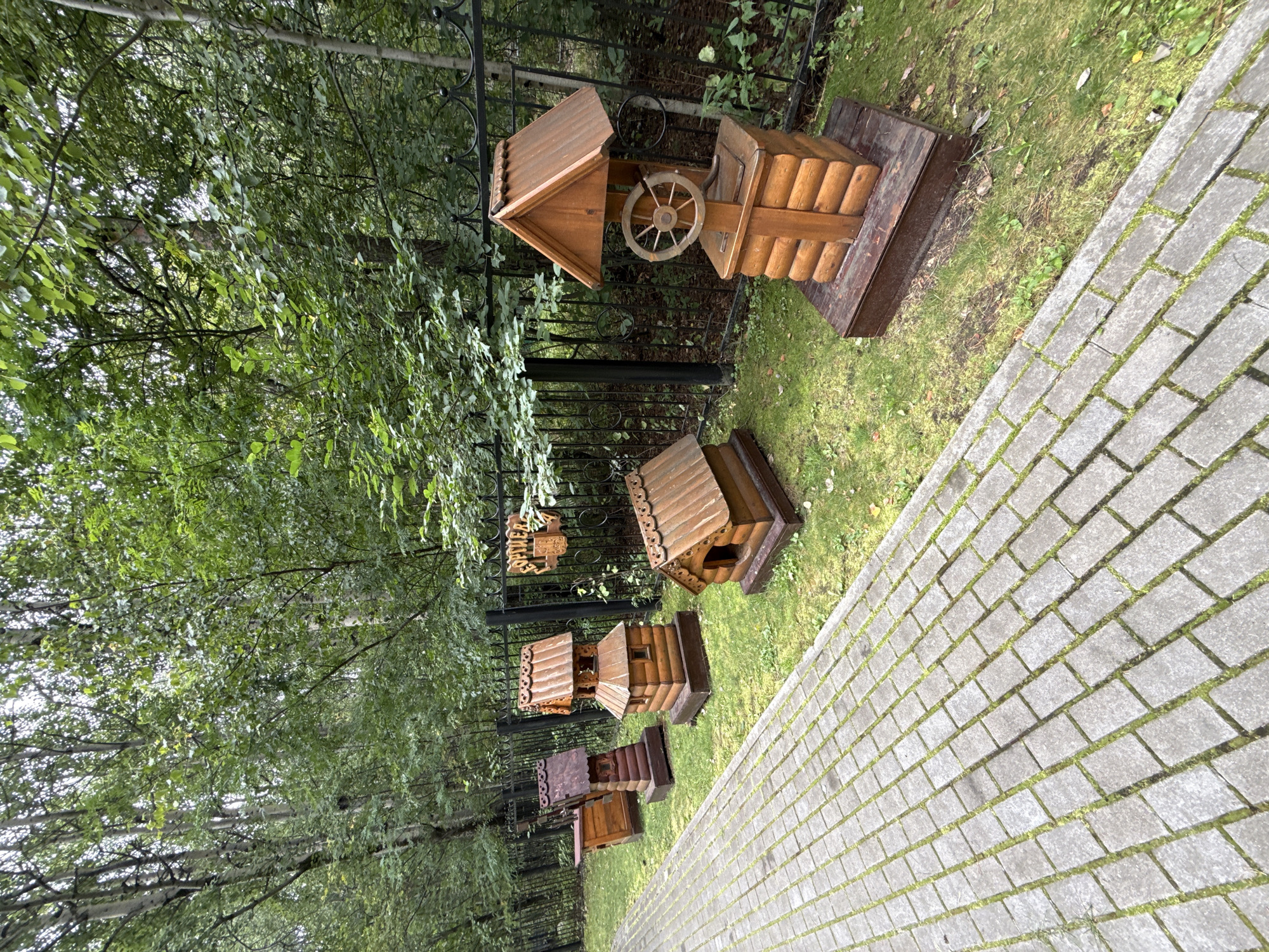 The image shows a cobblestone path leading to a row of decorative wooden wells and structures situated on a grassy area in front of a black metal fence. Trees and foliage are visible behind the fence. There are at least five wooden structures, including a well with a wheel, a smaller well with a pointed roof, a doghouse-like structure, and two other elongated wooden buildings. One of the structures has a sign with Cyrillic lettering. The scene appears to be outdoors in a park-like setting.