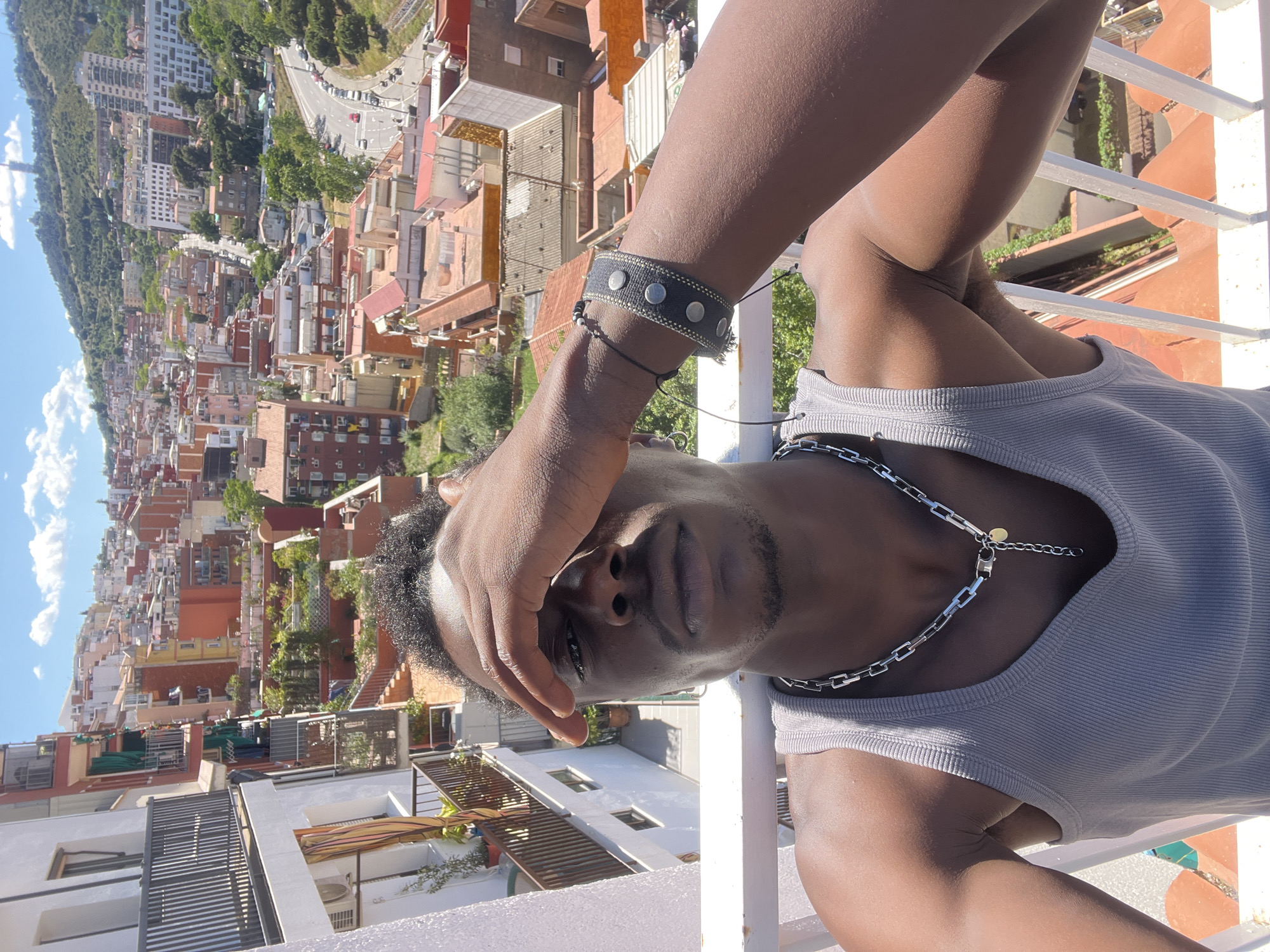 The image captures a man from a low angle on what appears to be a rooftop or balcony in Barcelona, Spain, looking directly at the camera. He covers part of his face with his hand, casting a shadow over his eyes. He has short hair, a mustache, and a goatee. The man is wearing a grey tank top and a silver chain necklace. A studded wristband is visible on his wrist. The background shows a densely packed urban landscape of Barcelona with orange and white buildings, a winding road leading up a hill, and trees. The sky is blue with a few clouds. The foreground includes the white railing of the balcony.