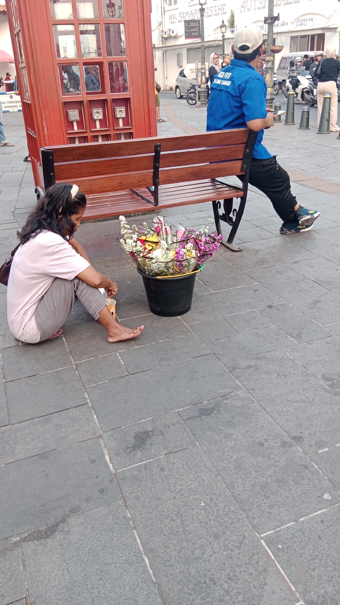 The image depicts a street scene in the Old City of Semarang, Indonesia, specifically in the Kota Lama area. In the foreground, a young woman with long, dark hair and a light pink shirt and grey pants is sitting on the paved ground, facing a black bucket filled with a colorful assortment of flowers. She appears to be arranging or preparing the flowers, holding a small cup or container in her right hand. To her right, and slightly behind her, is a red, replica British-style telephone booth. A wooden bench is positioned in front of the telephone booth, with a portion of its slats visible.
In the middle ground, a man wearing a blue t-shirt with text and black pants is sitting on the bench, facing away from the camera. He is wearing a white baseball cap and athletic shoes. His left arm is resting on the back of the bench. In the background, there are more people walking and standing, motorbikes parked, and buildings with signage, consistent with an urban environment. The ground is paved with large, grey stone tiles, and there are street lights visible. The overall scene suggests a public space, possibly a tourist area, where street vendors may operate.